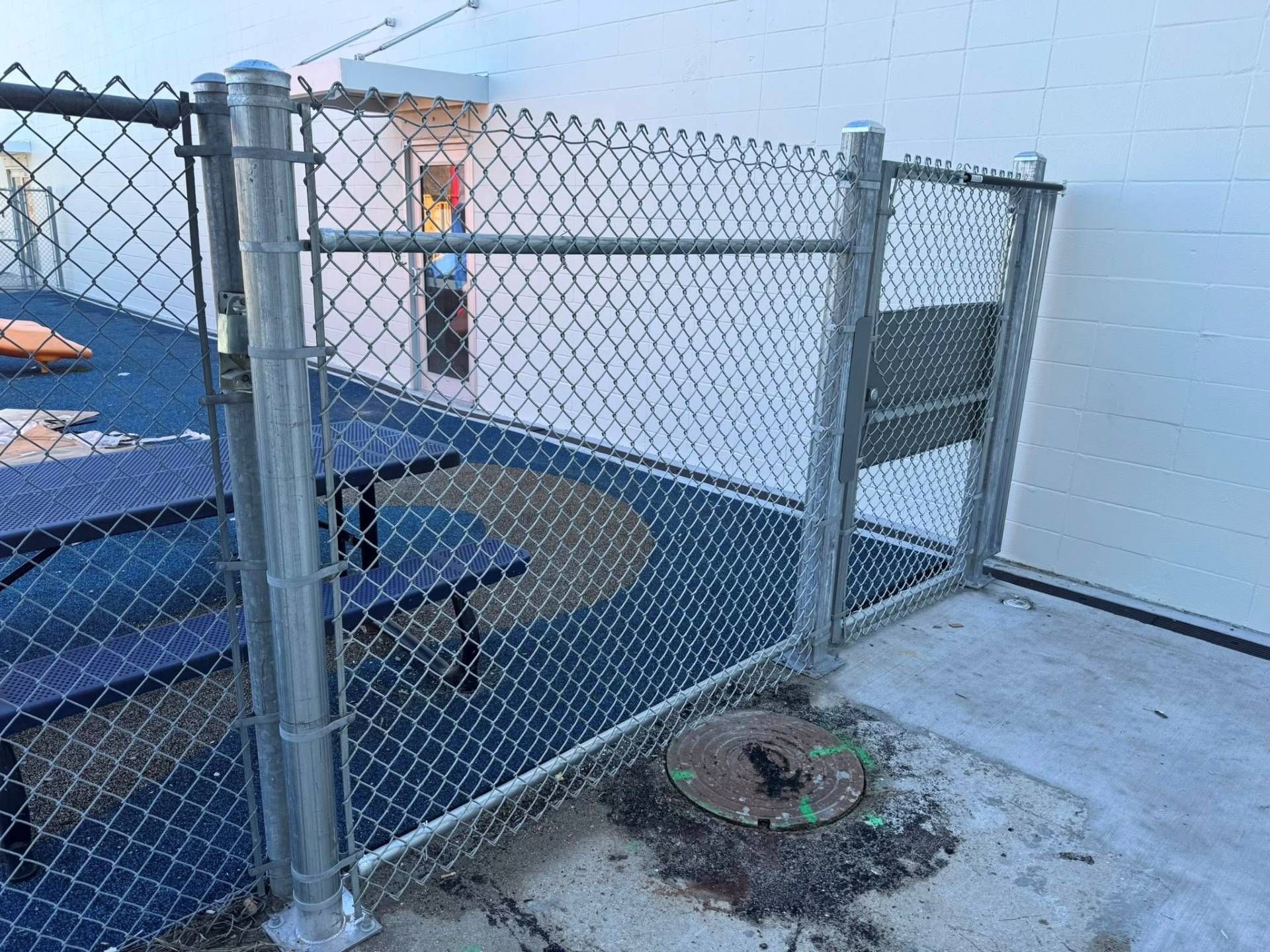 Chain link fence enclosing a playground area. Silver posts and gate, blue and brown play surface.