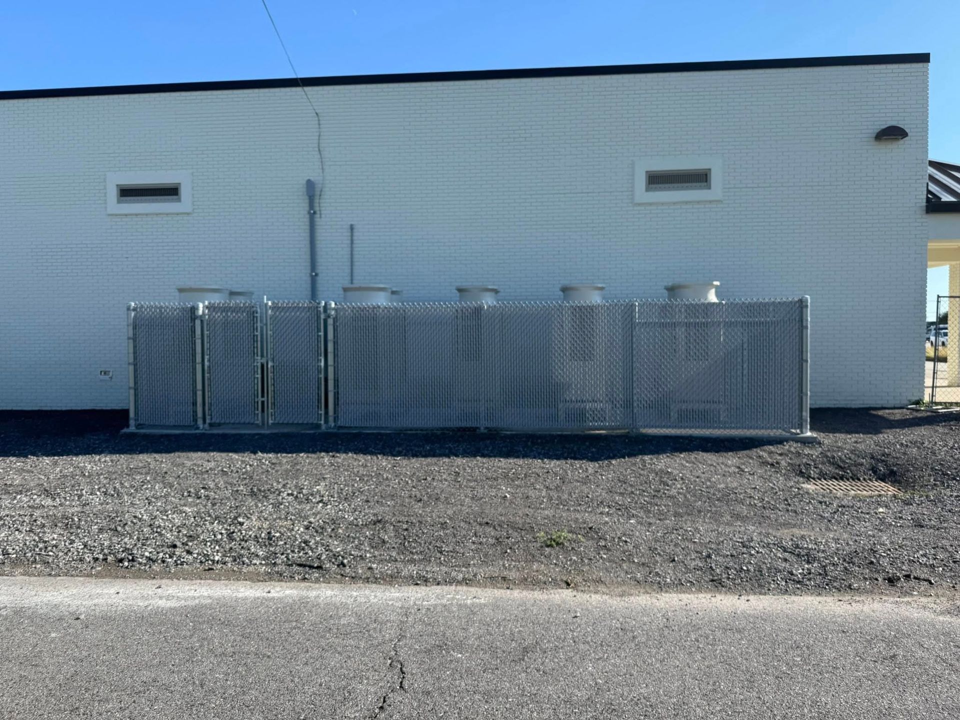Chain-link fence in front of a white building with air conditioning units behind it.
