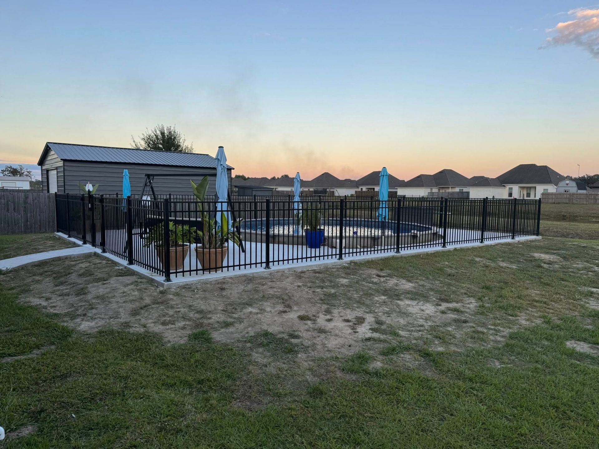 A backyard pool enclosed by a black fence, with blue umbrellas. Sunset in the background.