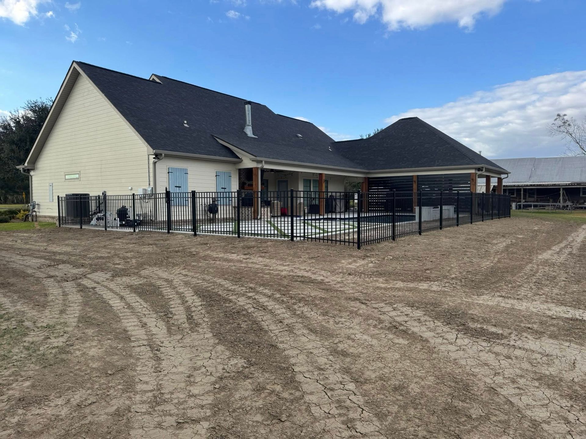 Backyard of a house with a black fence, pool, and dirt. Blue sky with clouds.