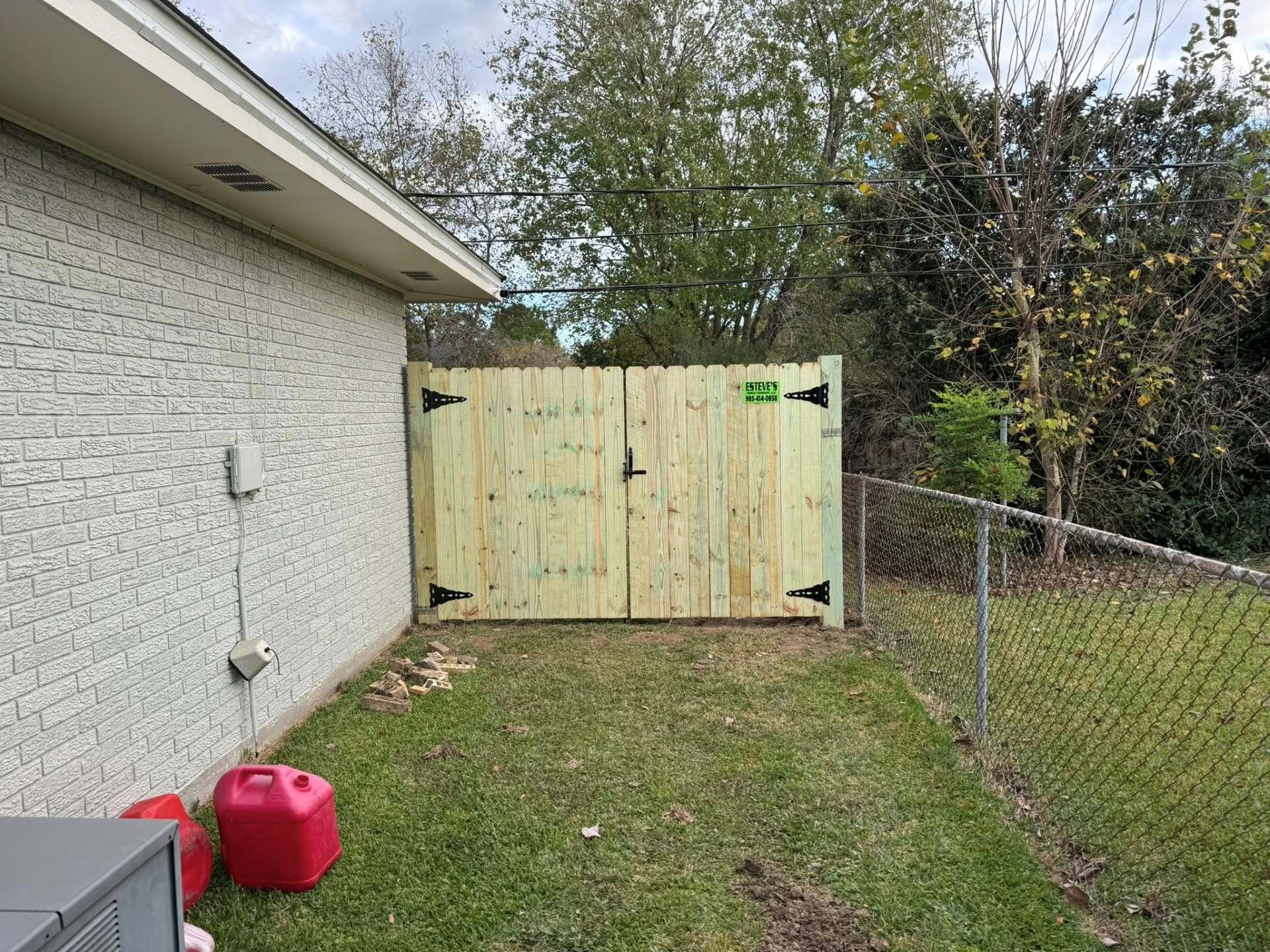 Wooden gate with black hinges and lock, set between a brick wall and a chain-link fence, on green grass.