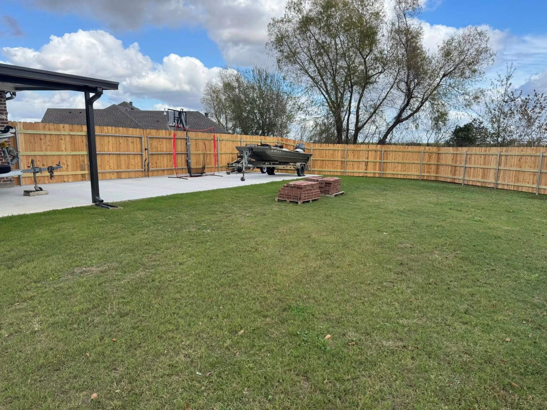 A backyard with a wooden fence, lawn, concrete patio, boat on a trailer, and a fire pit under a cloudy sky.