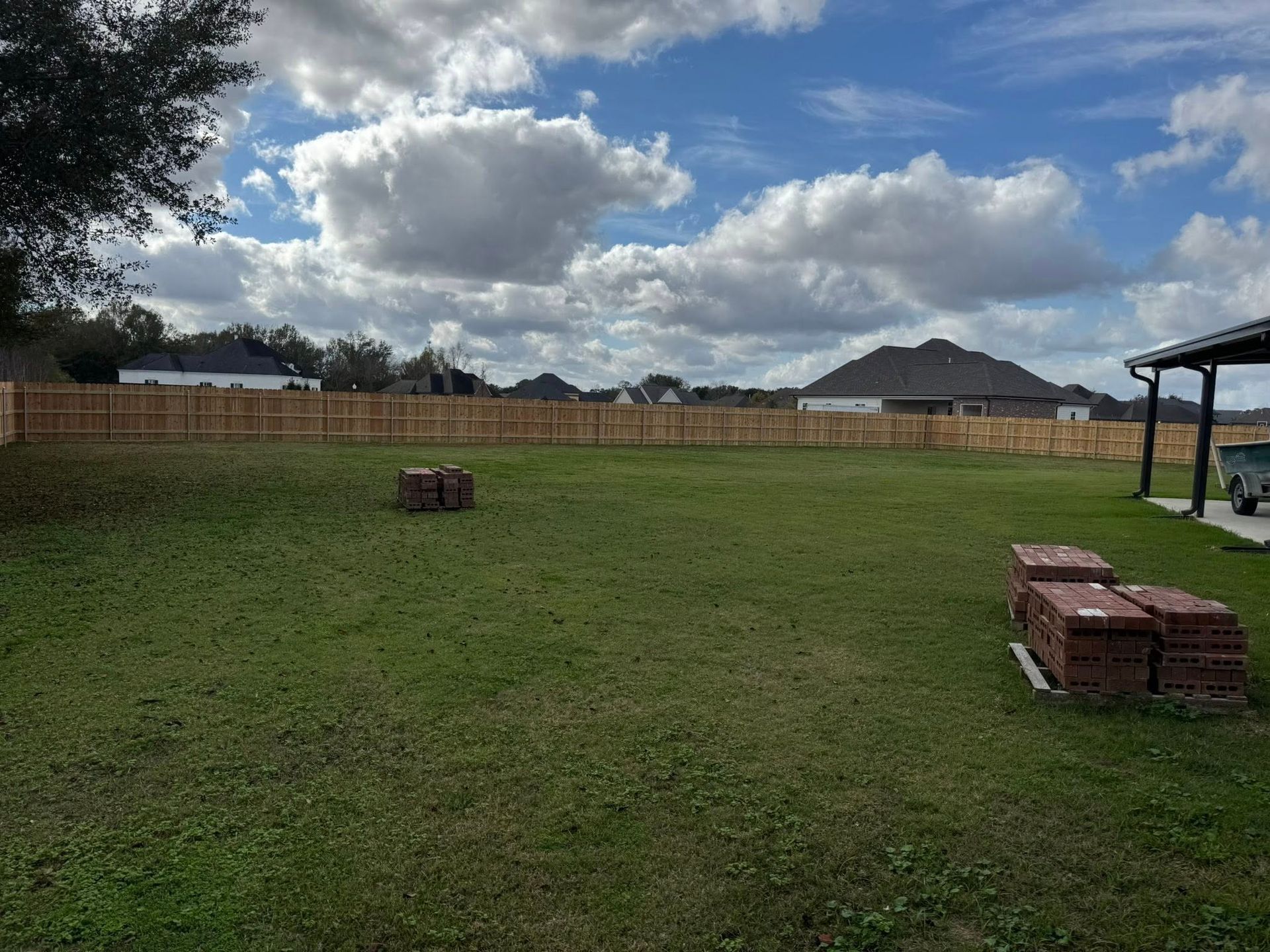 Grassy backyard with wooden fence, brick piles, and houses under a cloudy sky.