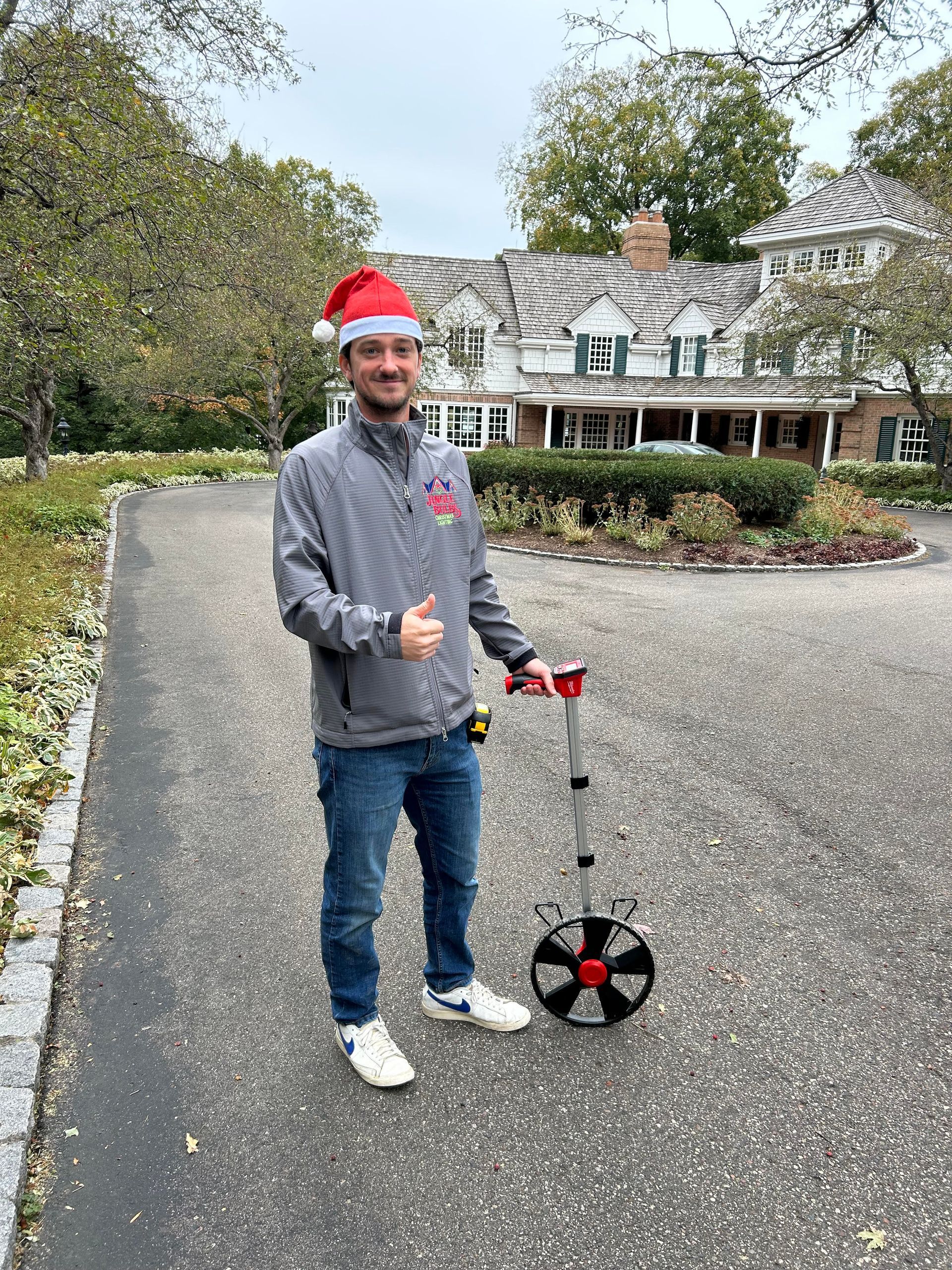 Man in Santa hat holds measuring wheel on driveway; large house in background.