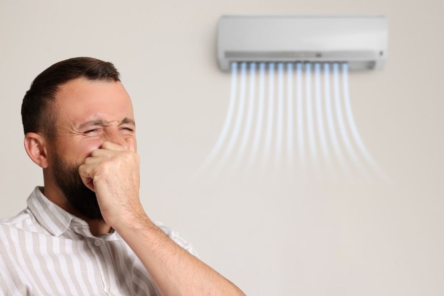 Homeowner covering his nose near a wall-mounted AC unit blowing musty air