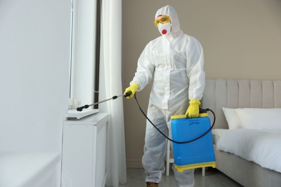 Technician in full white protective suit spraying antimicrobial treatment in a bedroom