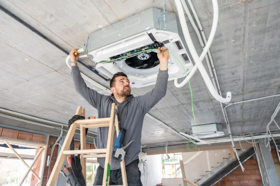 Technician on a ladder servicing an open ceiling-mounted HVAC cassette unit
