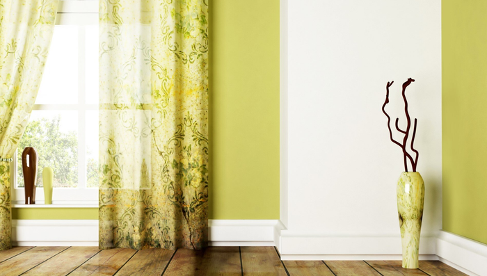 Lime green toned room with timber floor and tall case with wooden branch on floor. Green floral semi-opaque curtains with one side tied open, looking out over garden setting.
