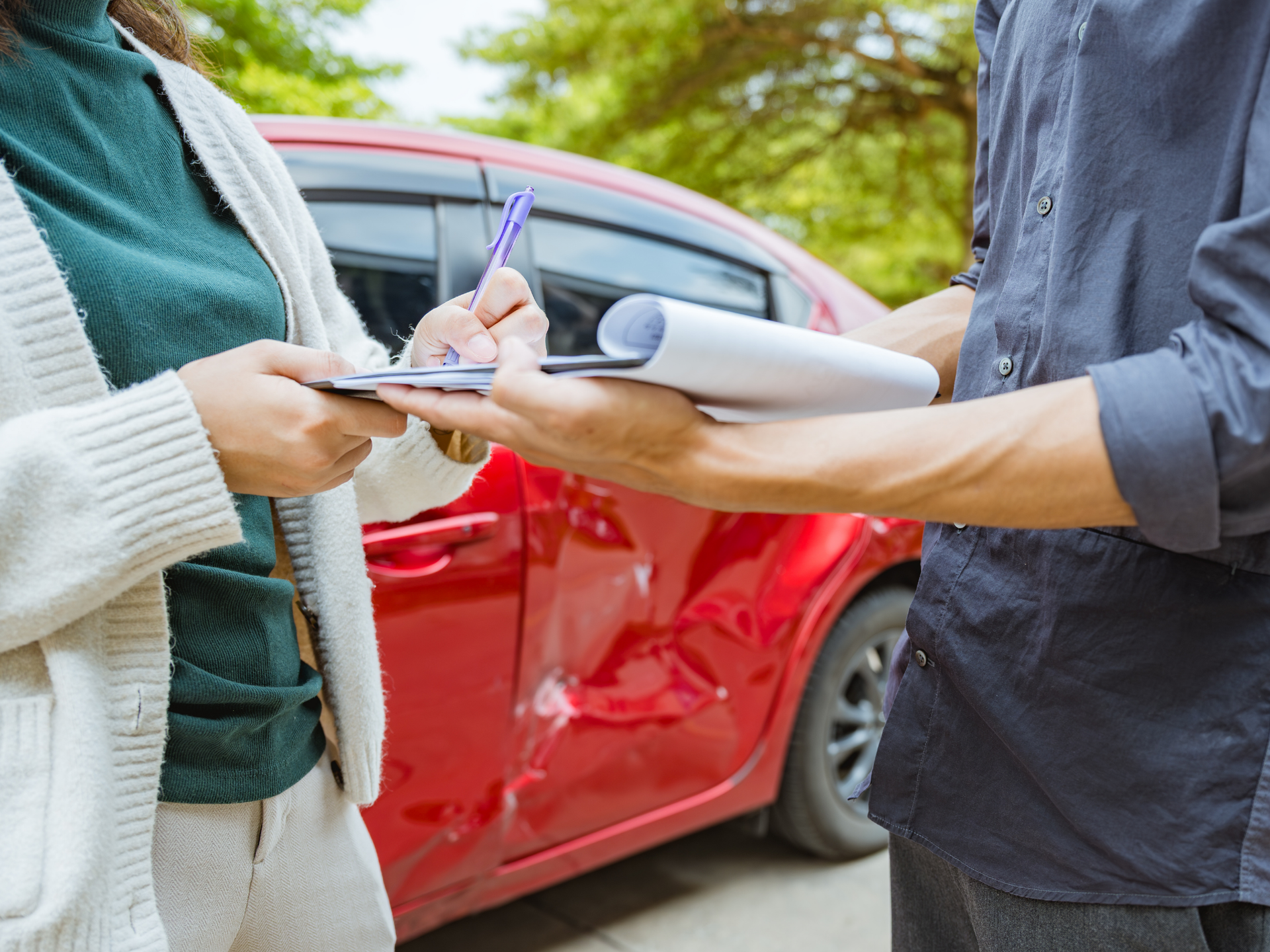 Person taking notes while inspecting a damaged red car, documenting a claim handled by a home and auto insurance broker service in San Tan Valley, AZ