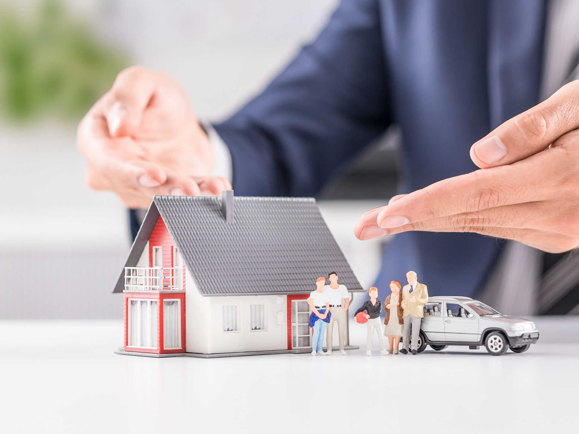 Hands placing a protective roof over a model house with a family and car, representing coverage from a home and auto insurance broker service in San Tan Valley, AZ