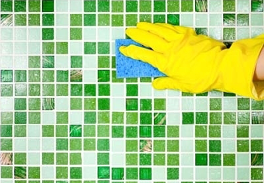 Una persona che indossa guanti gialli sta pulendo una parete piastrellata verde con una spugna.