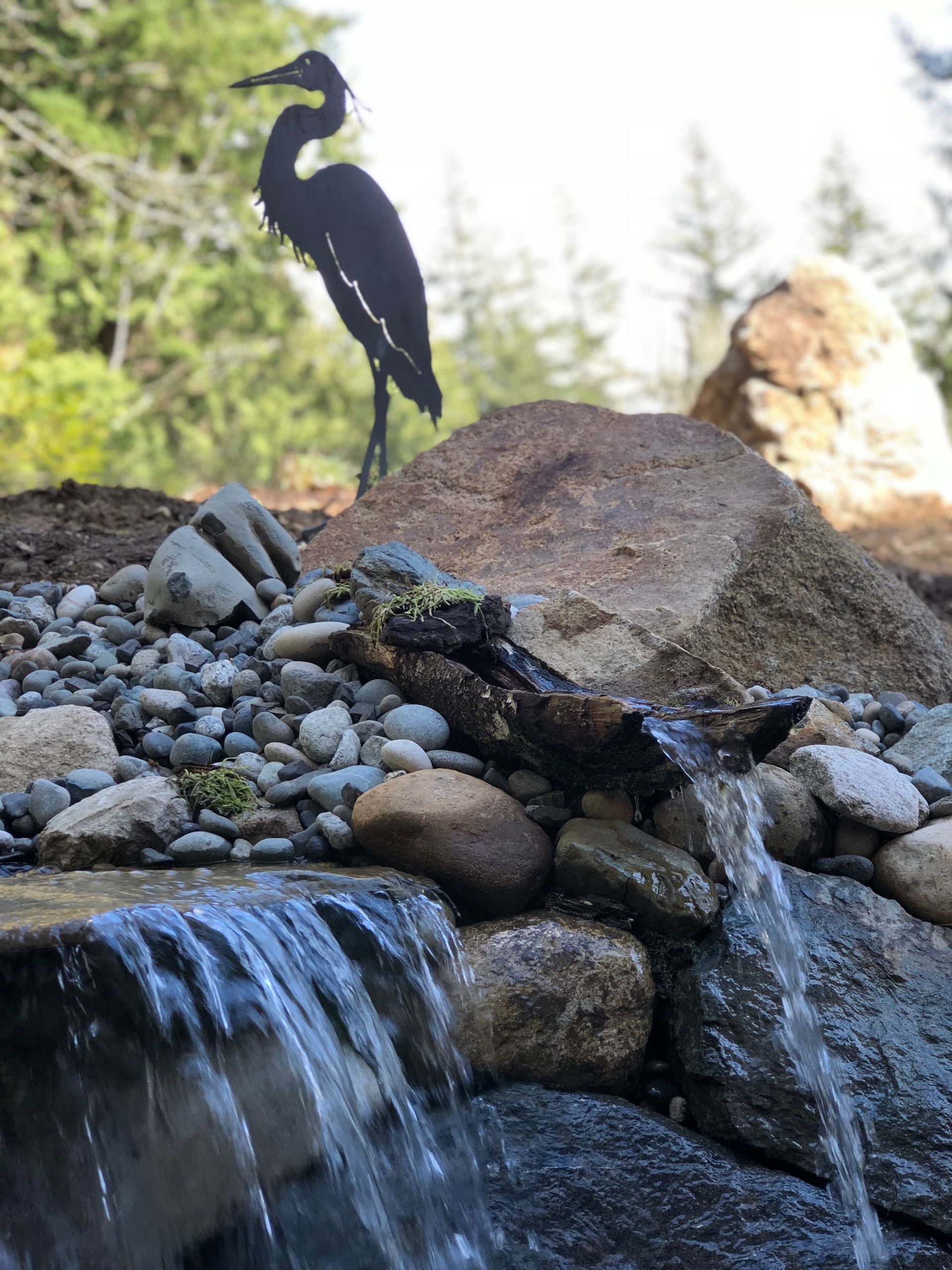 Black metal heron sculpture atop a rock formation by a waterfall; forest backdrop; work by West Coast Landscape & Hardscape. 