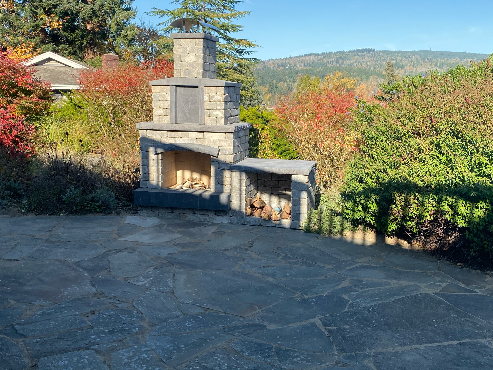 Stone outdoor fireplace with stacked design and wood storage, set on a patio with foliage and hills; work by West Coast Landscape & Hardscape. 