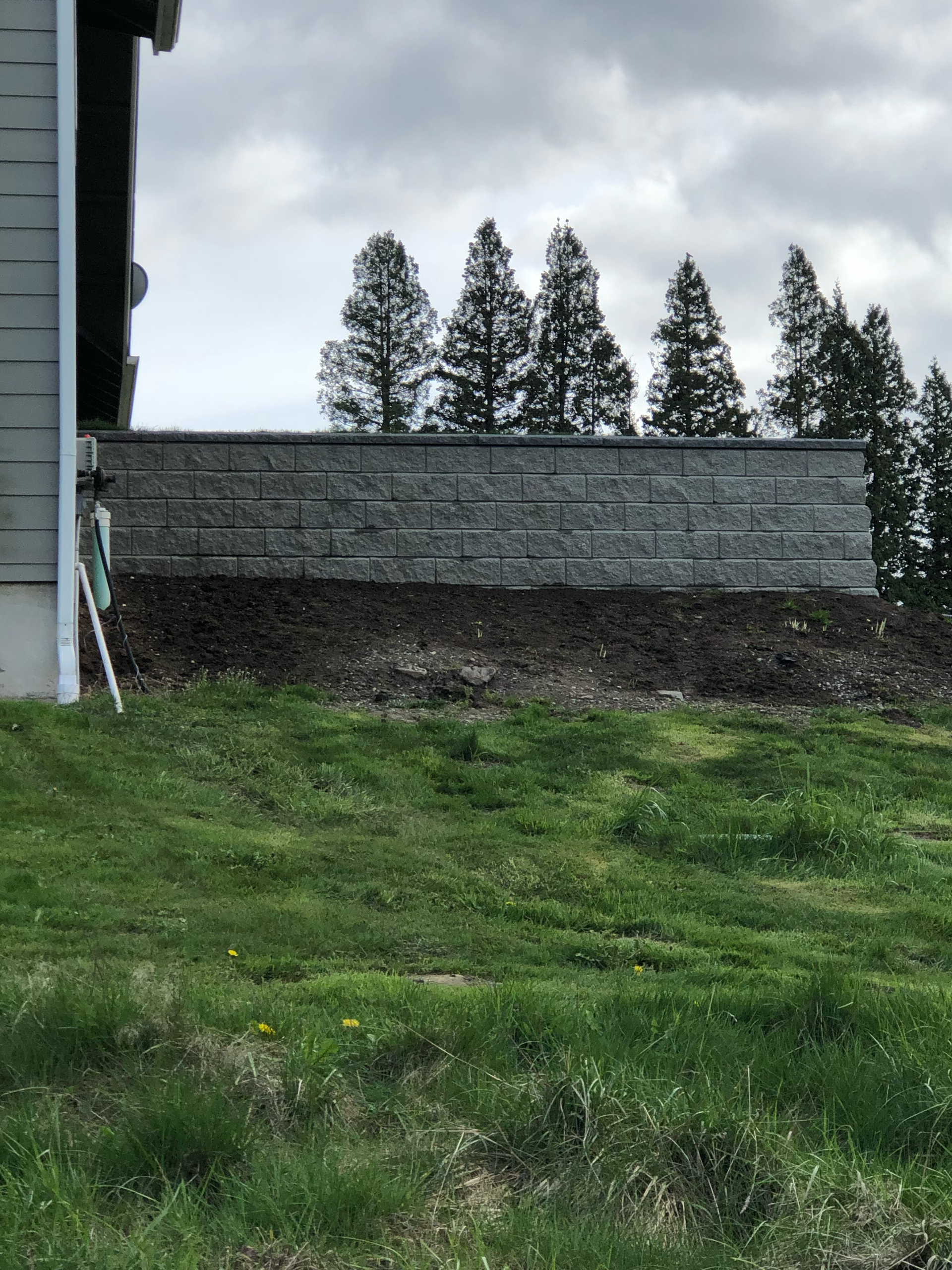 Gray retaining wall on a hill, with green grass in foreground and trees in background under cloudy sky; work by West Coast Landscape & Hardscape. 