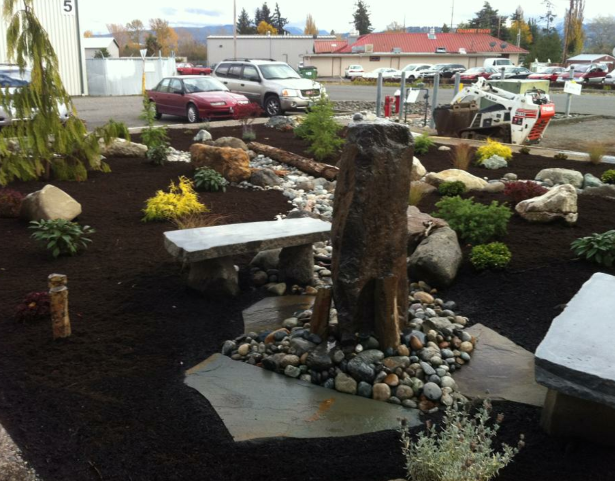 Landscaped area with water feature, stone bench, and various plants; cars in background; work by West Coast Landscape & Hardscape. 
