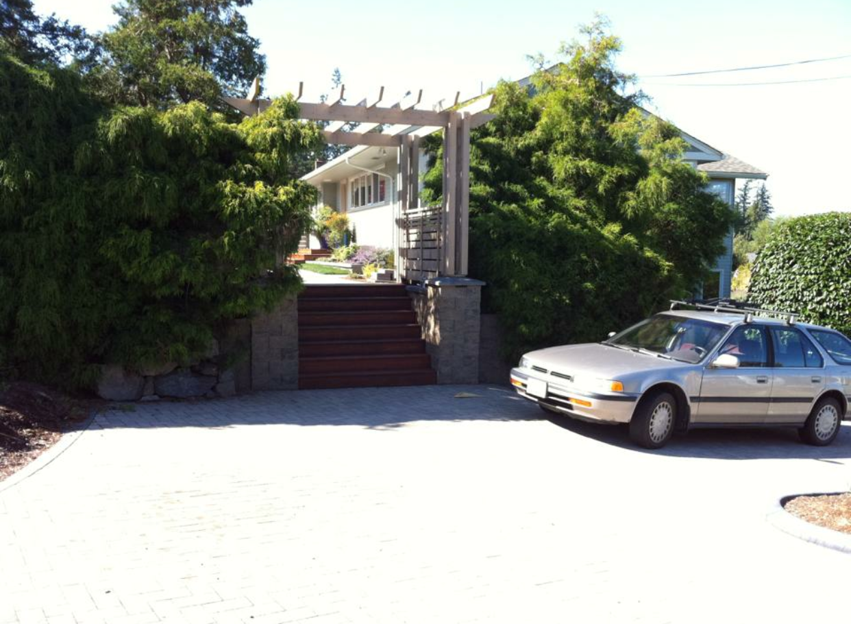 Silver car parked in front of a house with a wooden pergola entrance. Green shrubs flank the driveway; work by West Coast Landscape & Hardscape. 