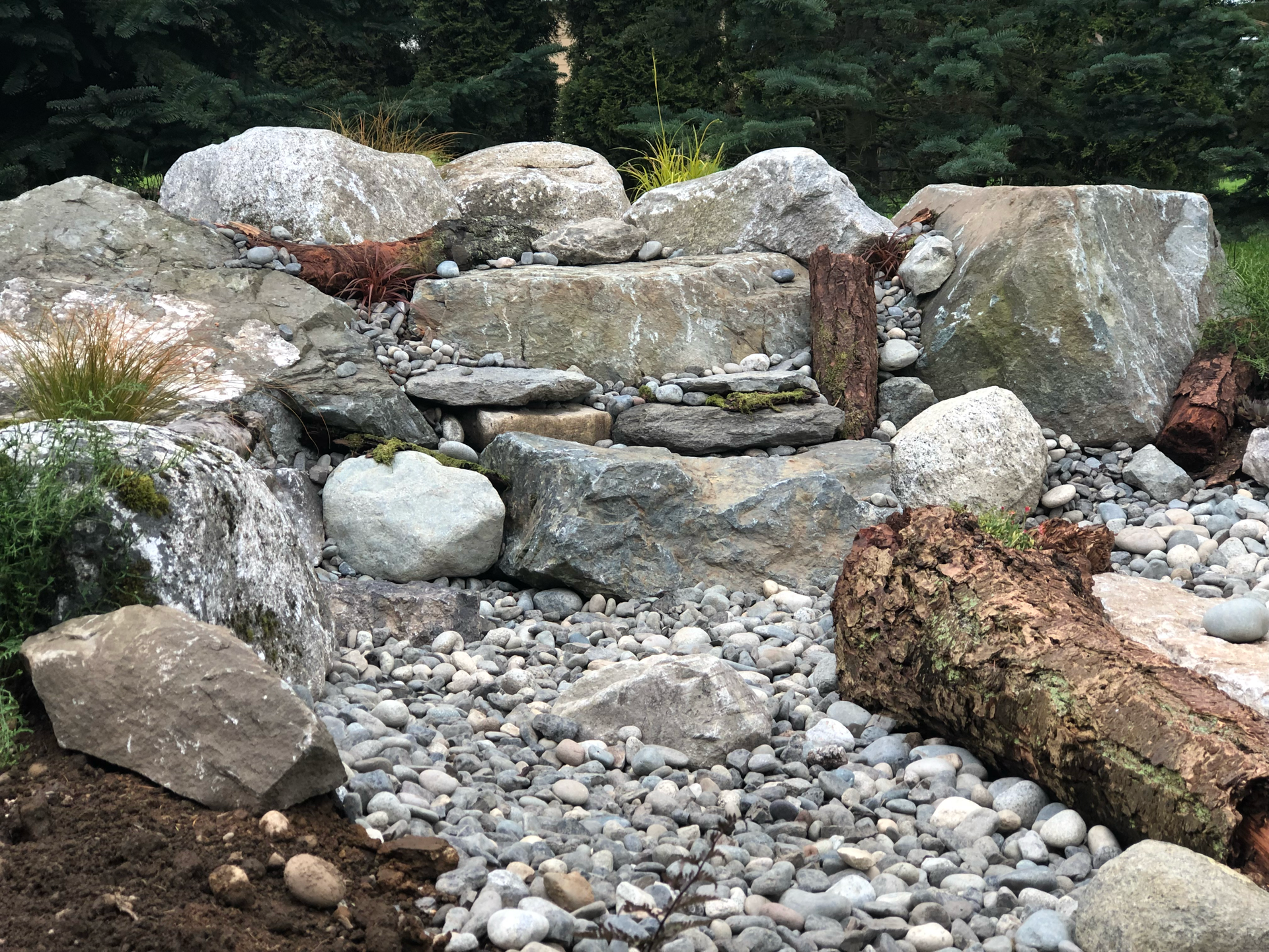 Rock garden with waterfall feature, gray rocks, pebbles, and a log in a natural outdoor setting; work by West Coast Landscape & Hardscape. 
