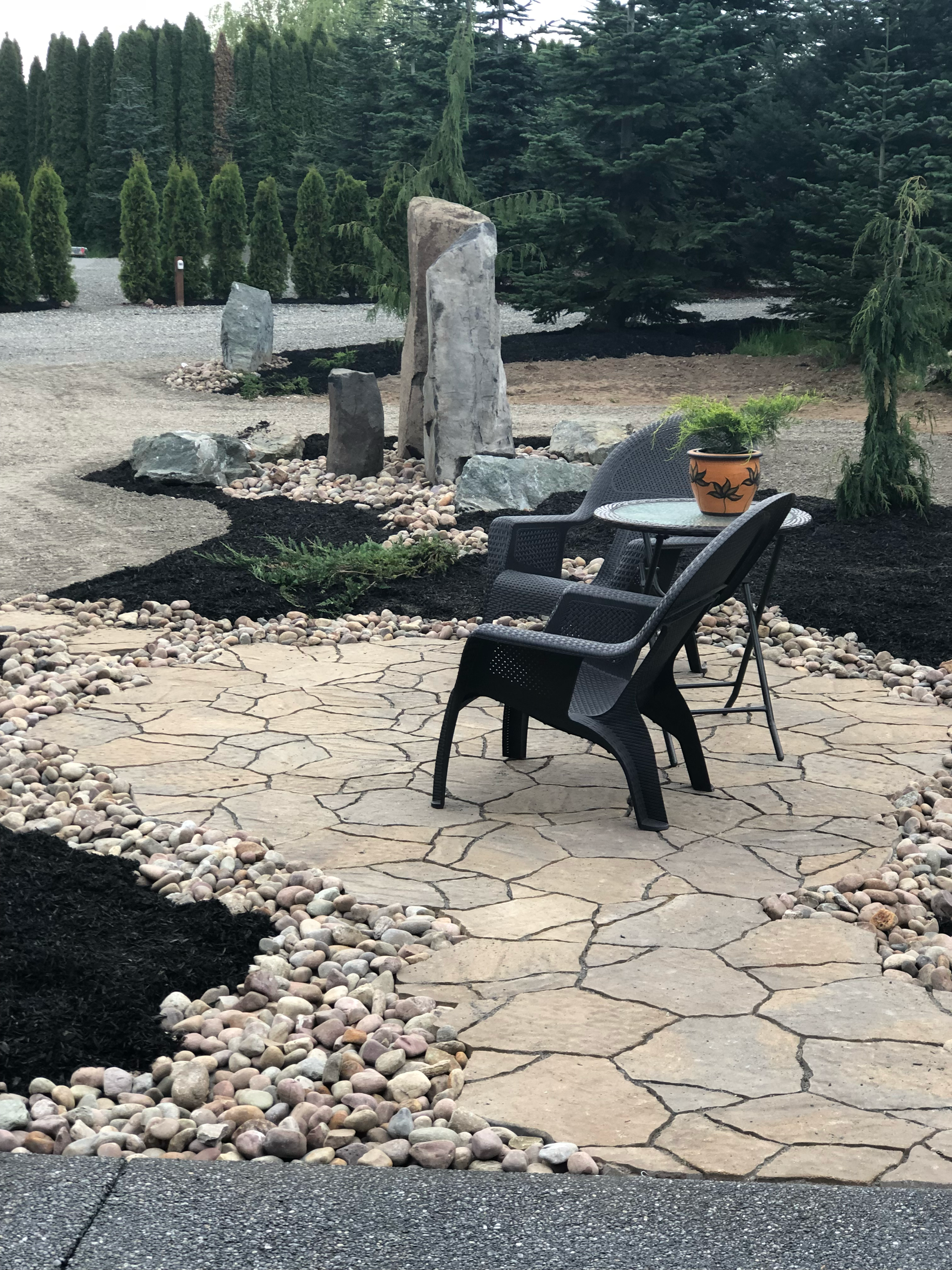 Stone patio with two black chairs, rock border, and large standing stone; work by West Coast Landscape & Hardscape. 