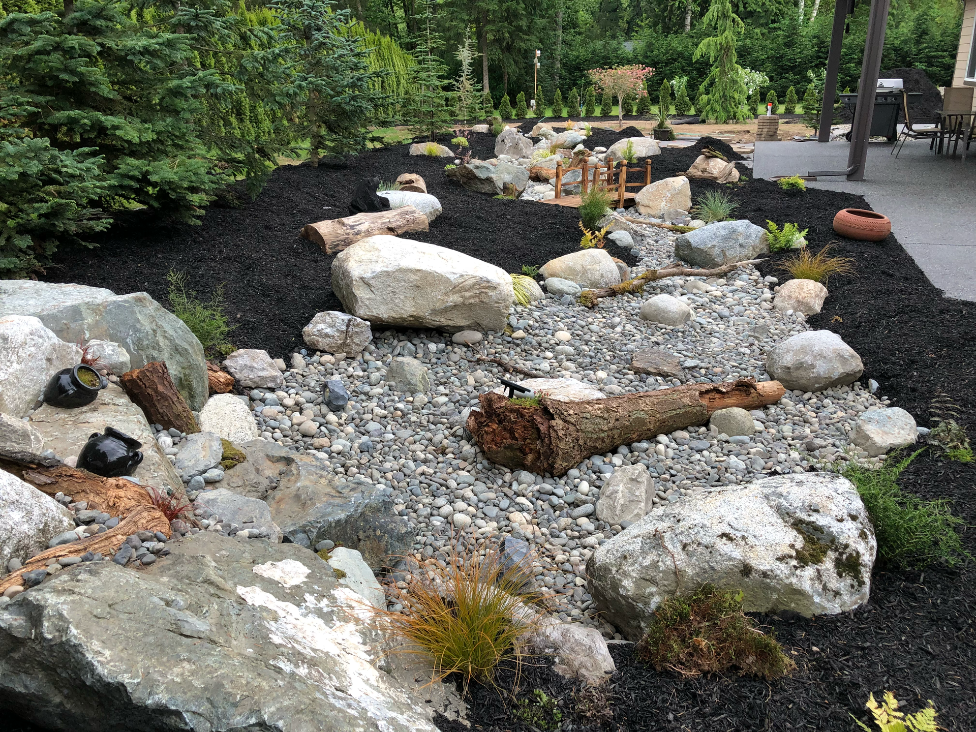 Rock garden with riverbed, rocks, logs, and dark mulch; work by West Coast Landscape & Hardscape. 
