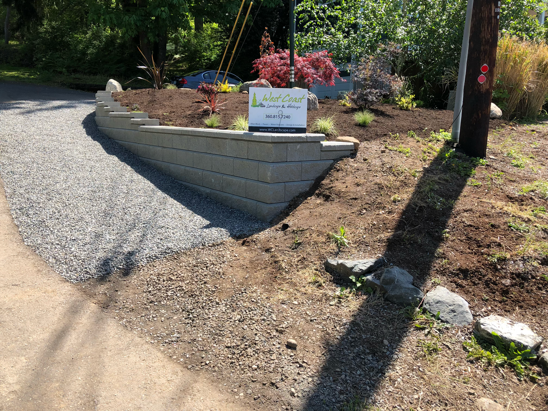 A gray retaining wall, gravel driveway and a sign with a landscaped garden; work by West Coast Landscape & Hardscape. 
