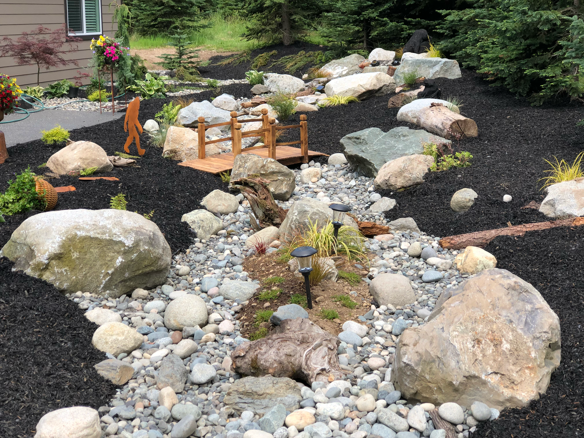 Landscaped stream with rocks, pebbles, a small wooden bridge, and black mulch; work by West Coast Landscape & Hardscape. 