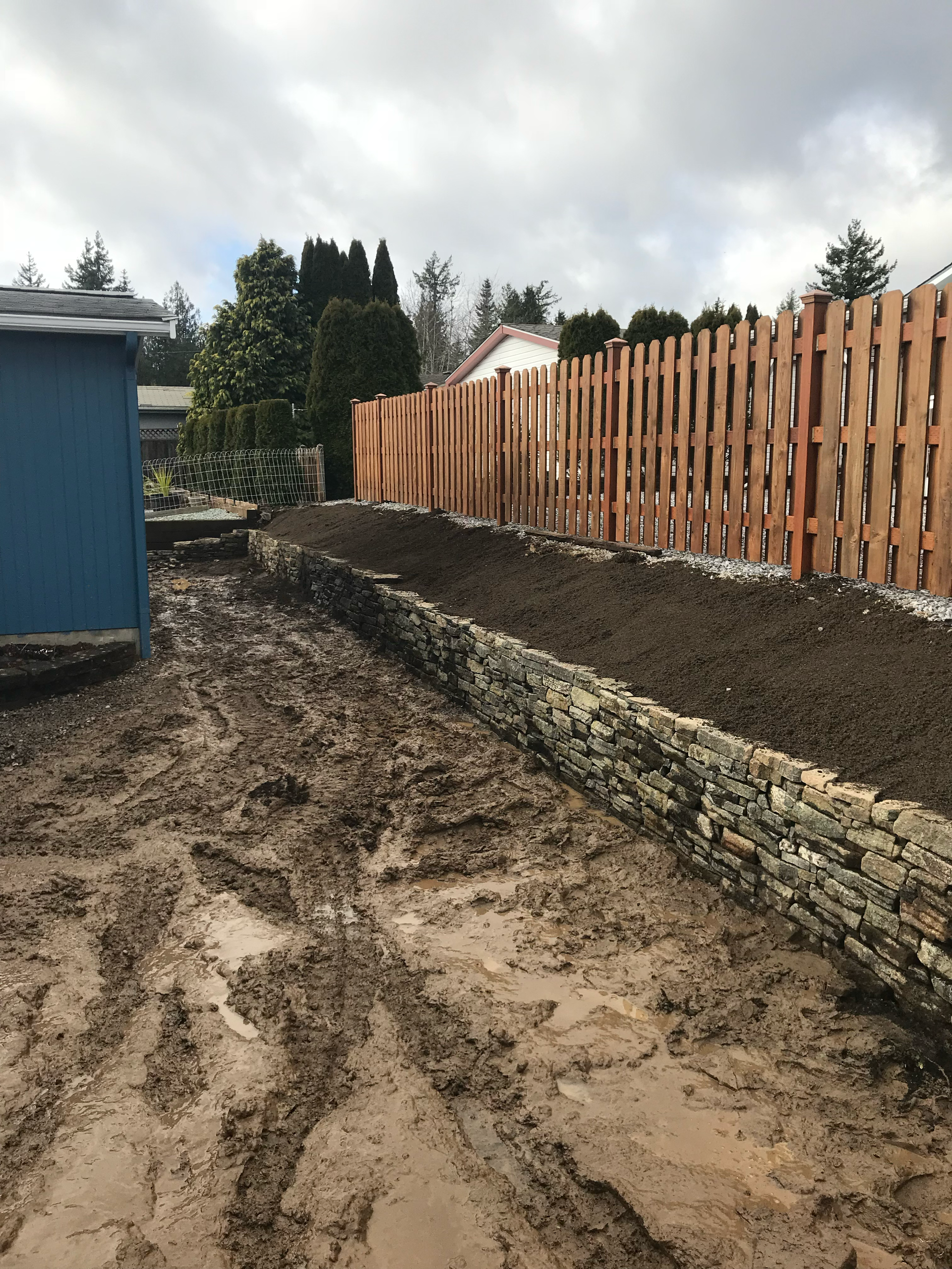 Muddy yard with gabion retaining wall, fresh soil, and wooden fence; work by West Coast Landscape & Hardscape. 