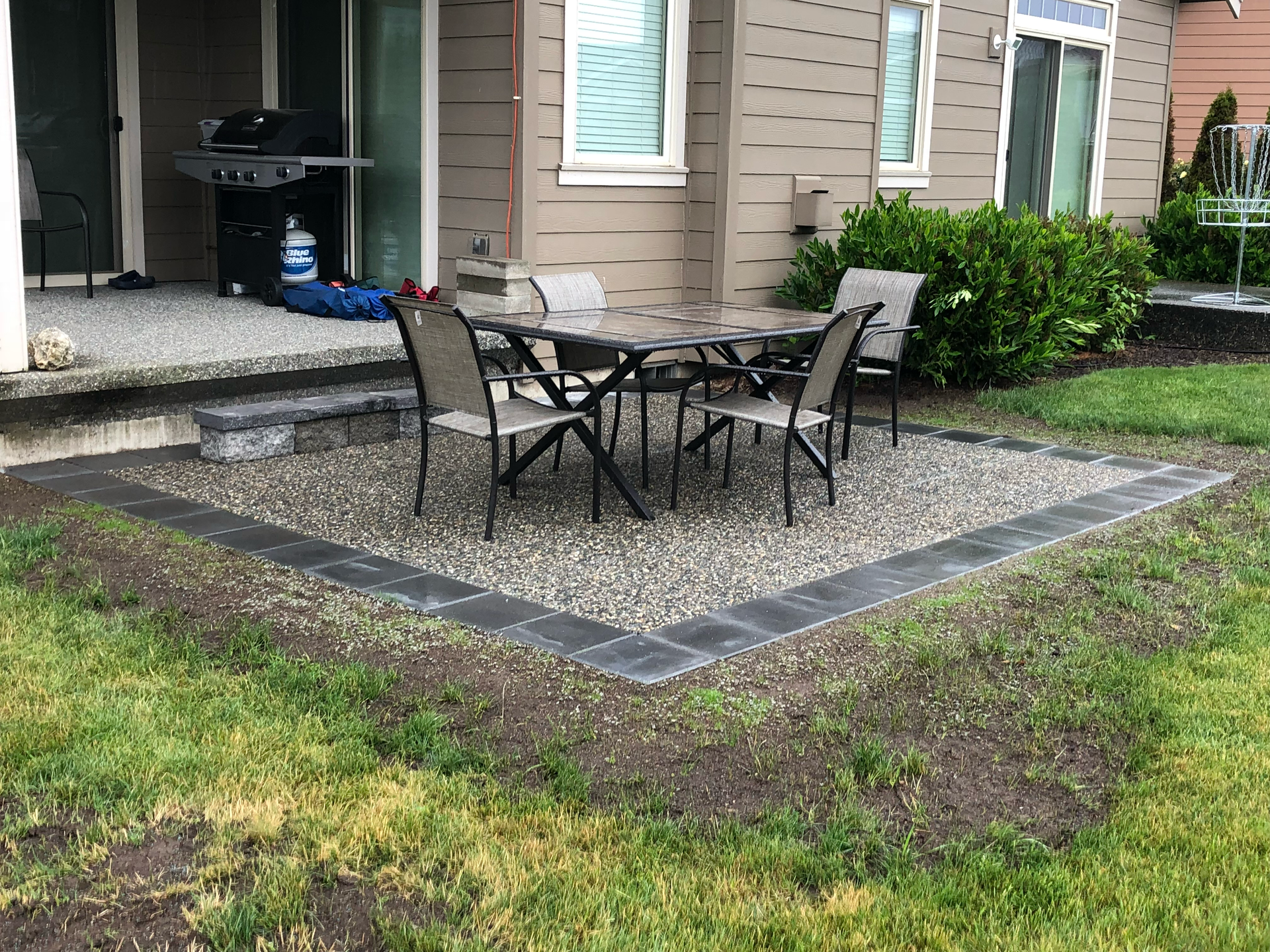 Patio with gravel and dark border, table with chairs on patio, next to house and grass; work by West Coast Landscape & Hardscape. 