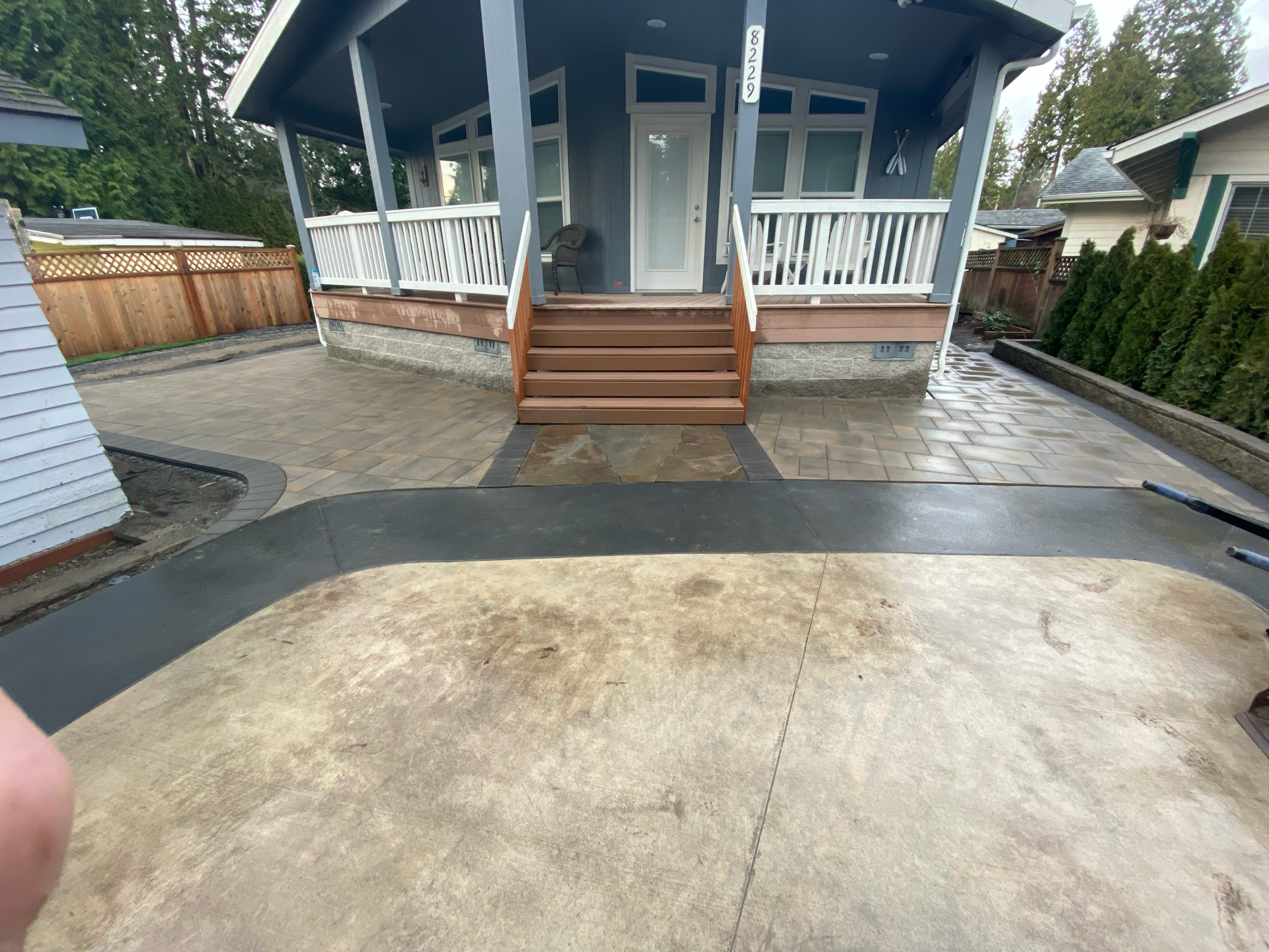 A small blue house with a porch and wooden steps leading to the front door. Grey concrete driveway; work by West Coast Landscape & Hardscape. 