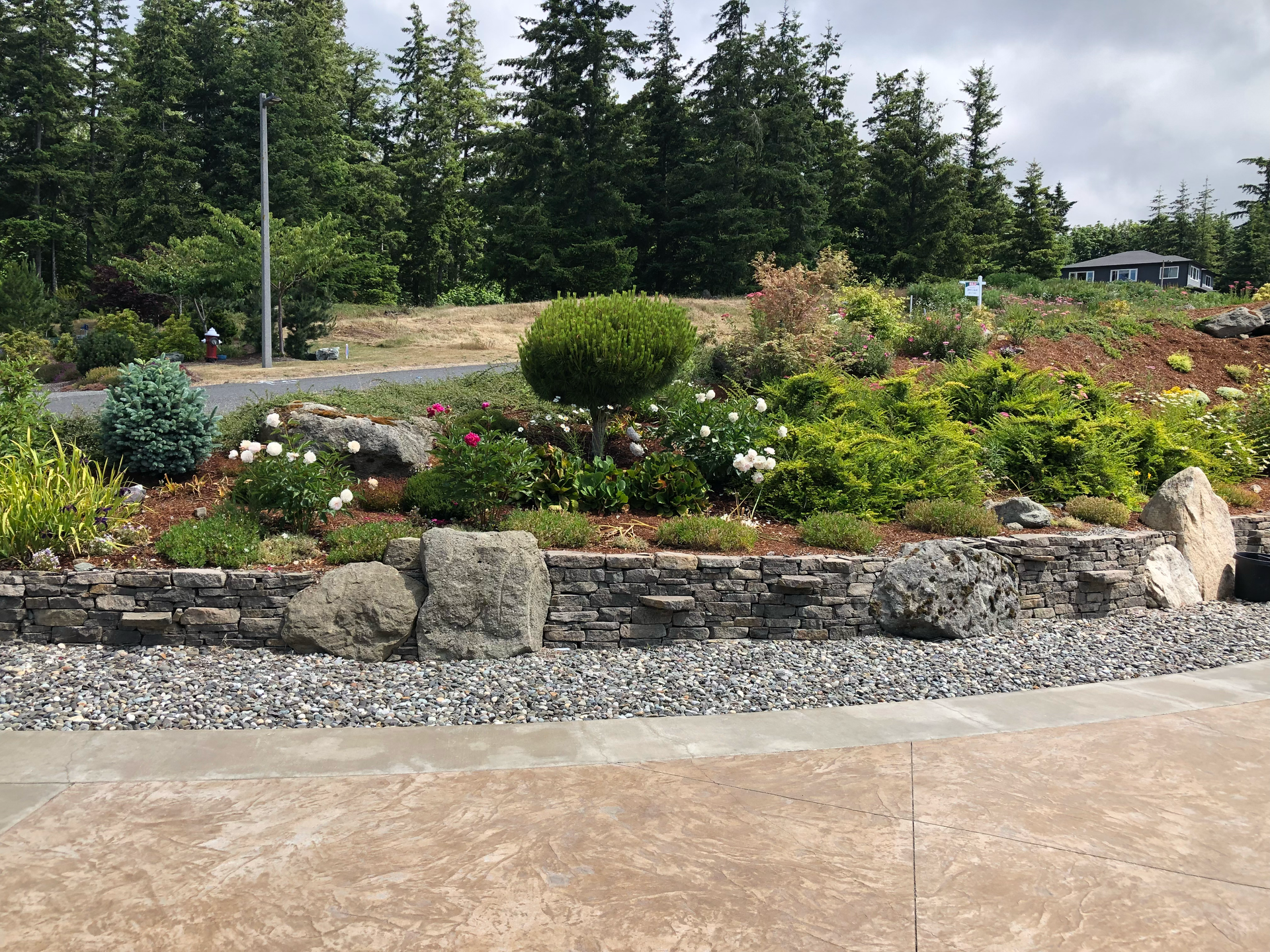 Stone retaining wall with a variety of colorful plants and shrubs, gravel, and a concrete path; work by West Coast Landscape & Hardscape. 