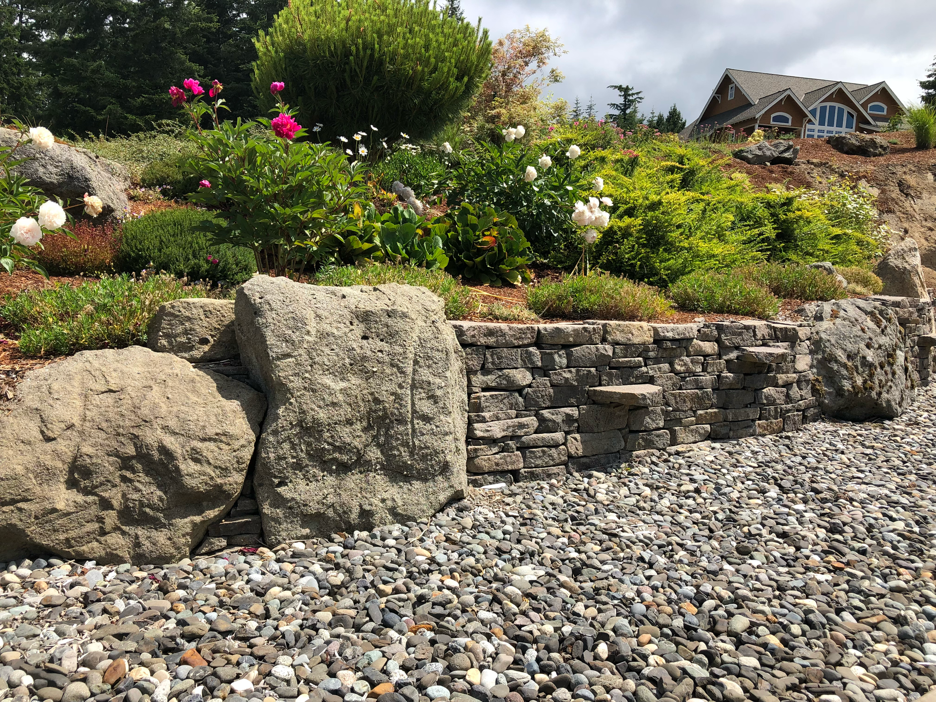 Stone retaining wall with colorful flowers and shrubs, leading to a house in the background; work by West Coast Landscape & Hardscape. 