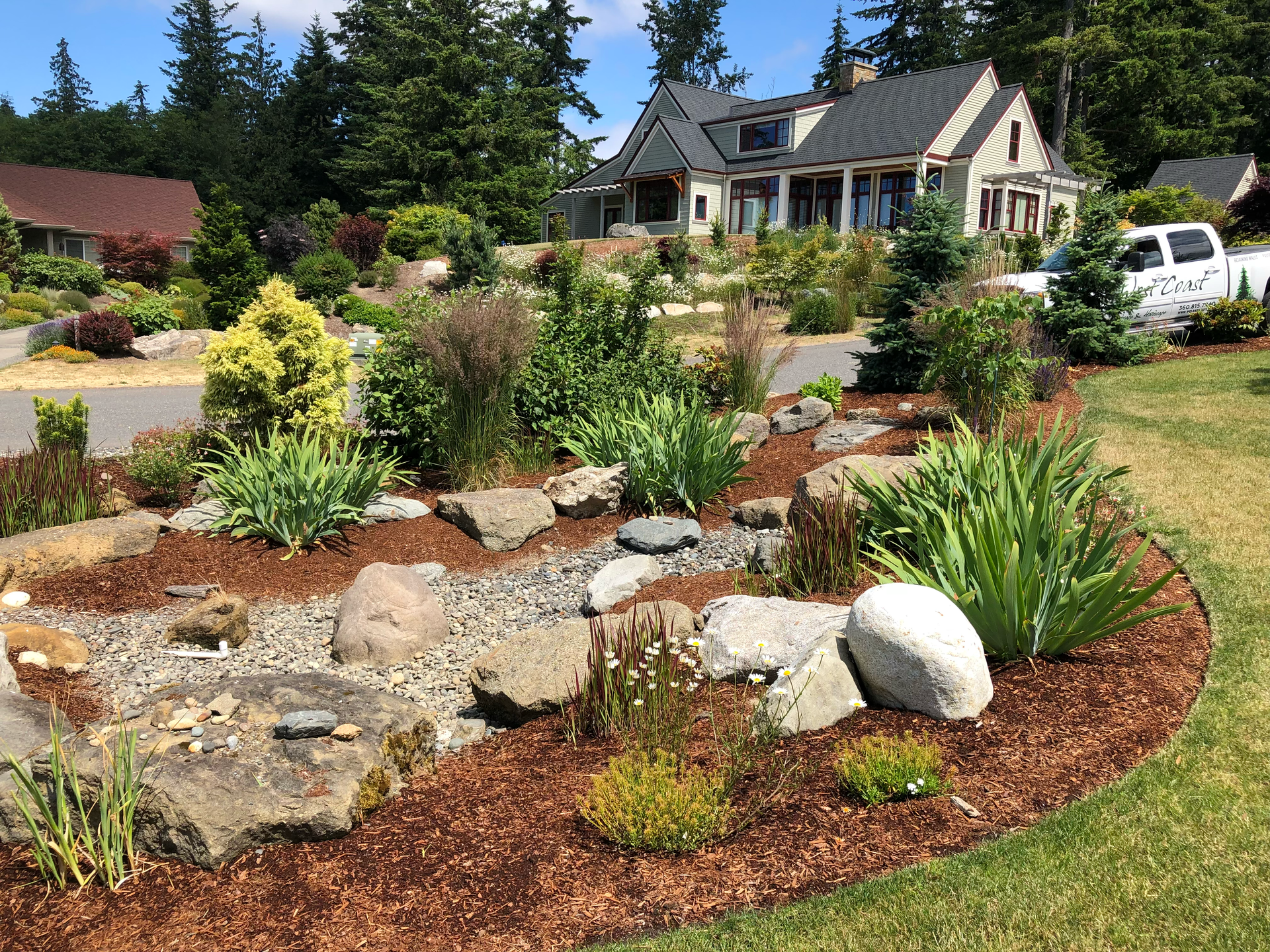 Landscaped yard with rock garden, brown mulch, and various plants in front of a house; work by West Coast Landscape & Hardscape. 