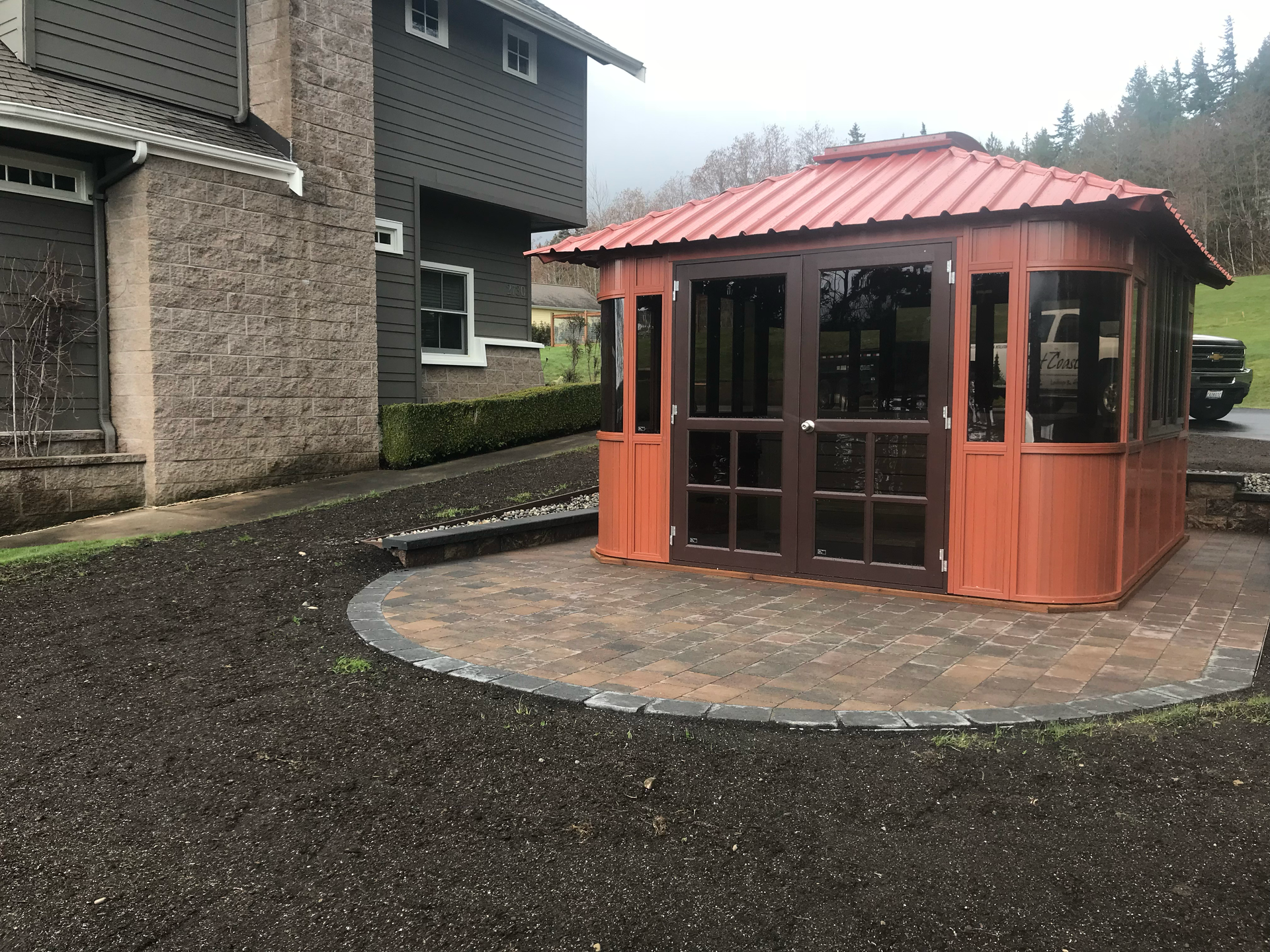 Orange gazebo with brown trim, set on a circular brick patio, next to a house with gray siding; work by West Coast Landscape & Hardscape. 