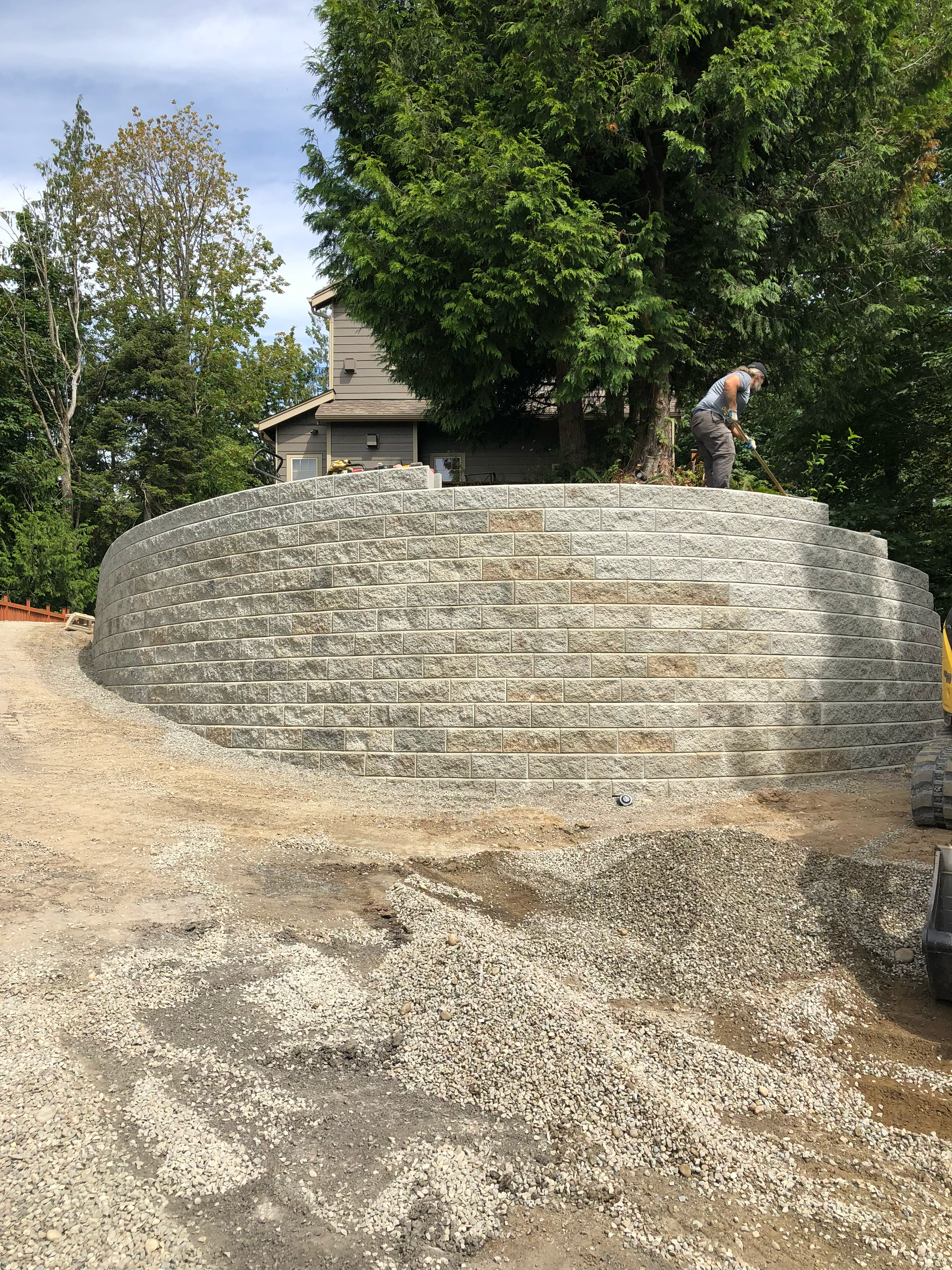 Stone wall construction in progress; person working on top of the curved structure near trees and gravel; work by West Coast Landscape & Hardscape. 