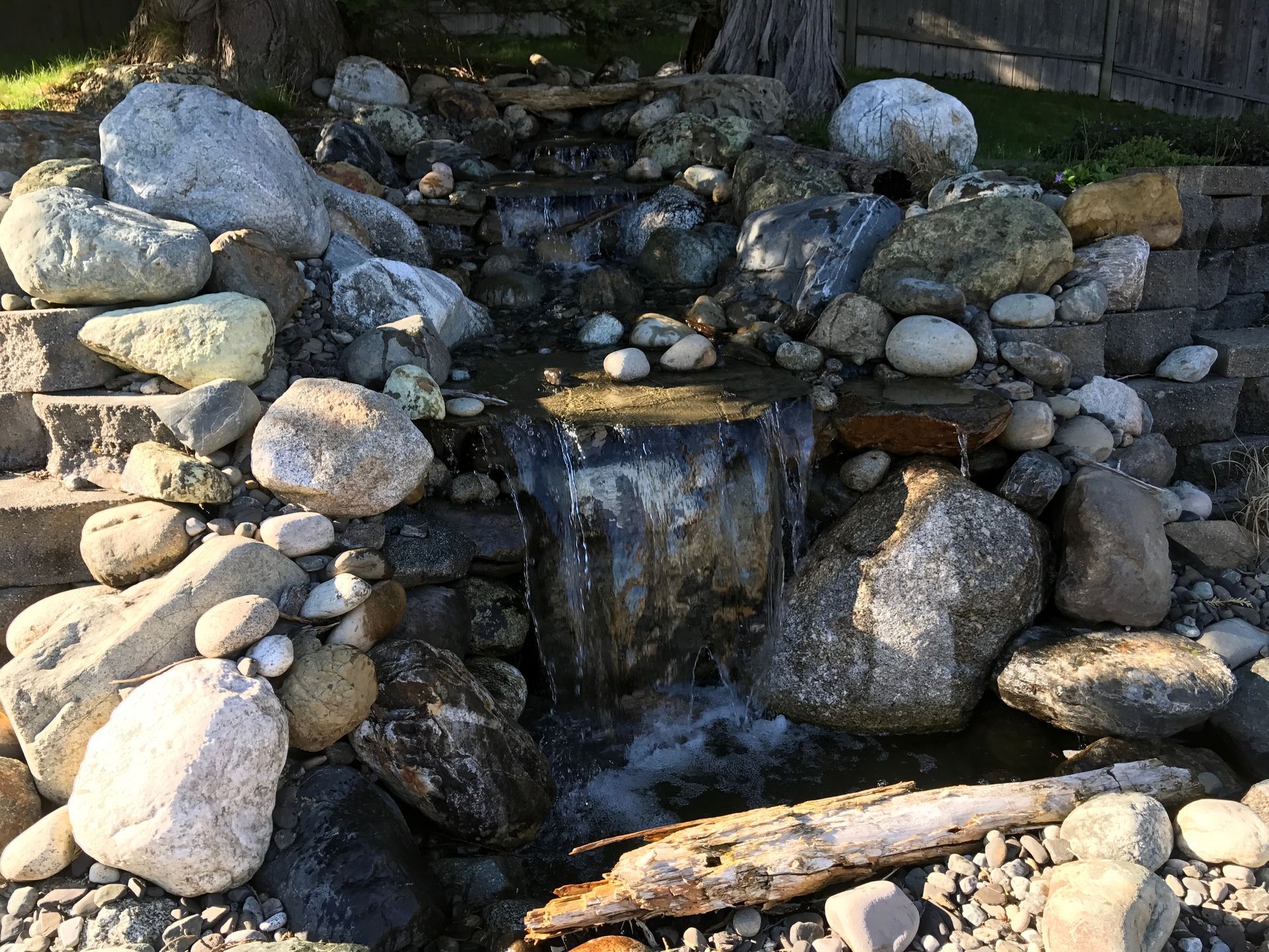 Small waterfall cascading over rocks and stones in a garden setting; work by West Coast Landscape & Hardscape. 