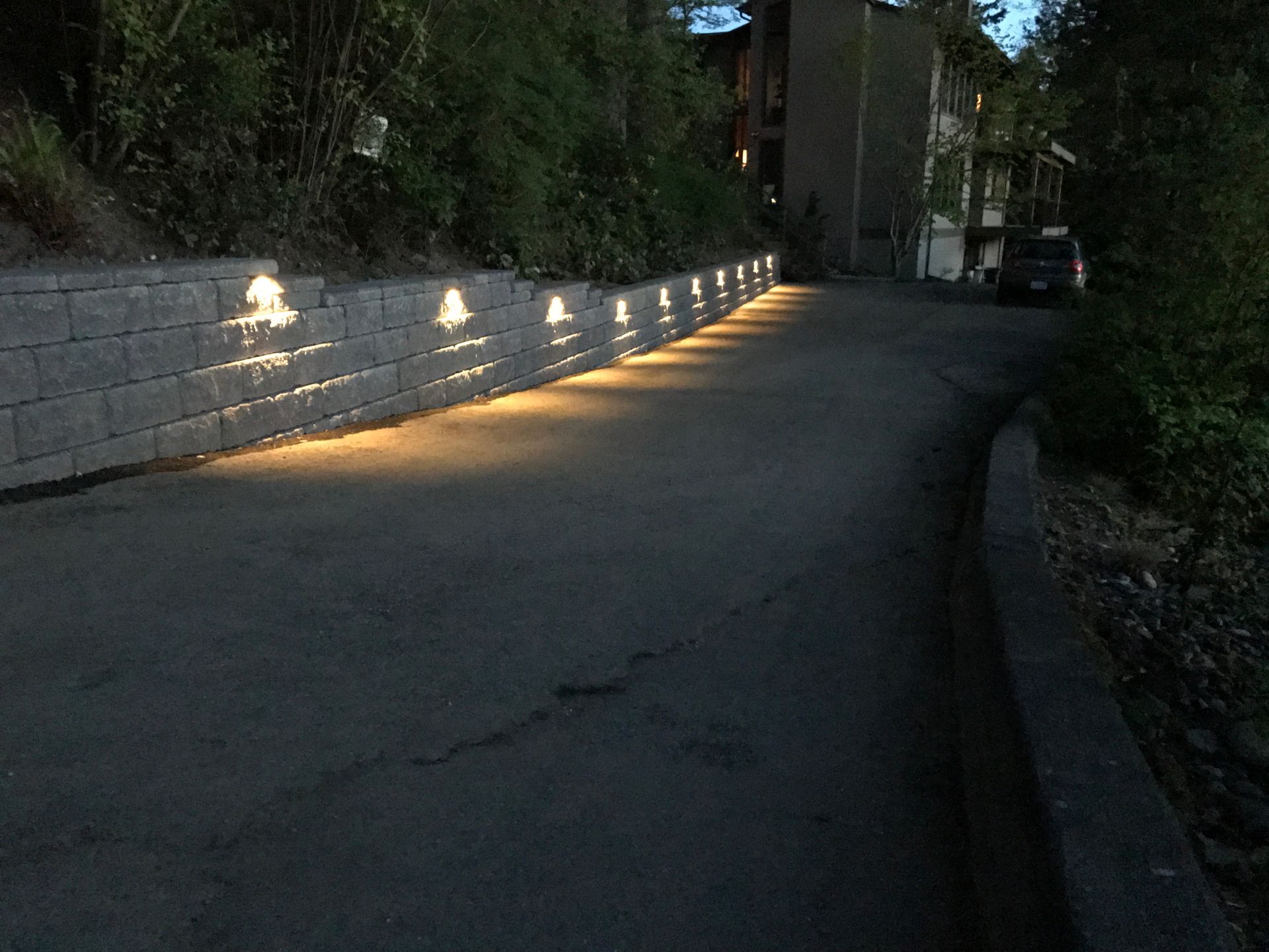 A dark asphalt driveway illuminated by wall-mounted lights along a retaining wall; work by West Coast Landscape & Hardscape. 