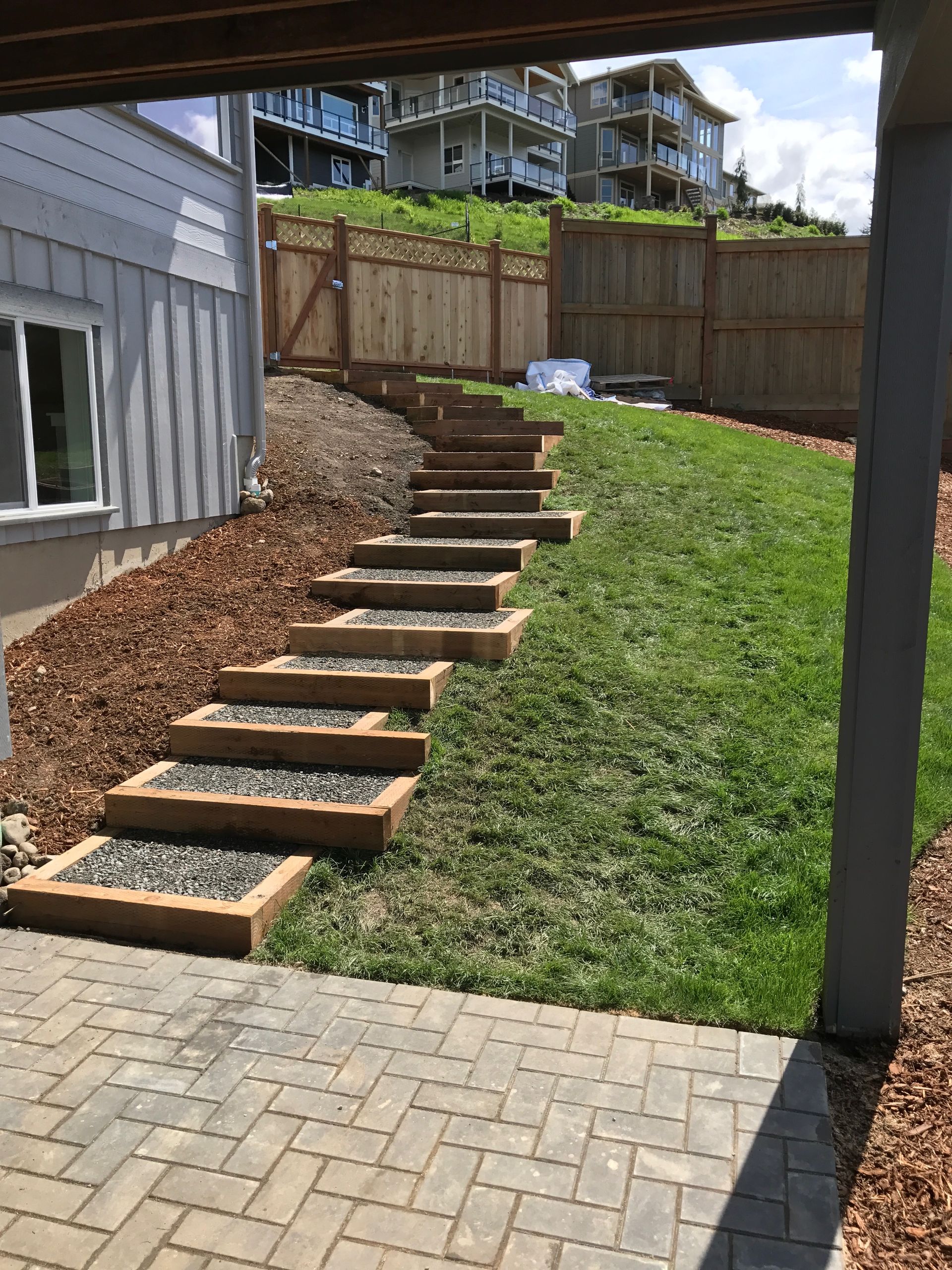 Stone steps with gravel and wooden borders ascend a grassy slope. Brick patio in foreground; work by West Coast Landscape & Hardscape. 