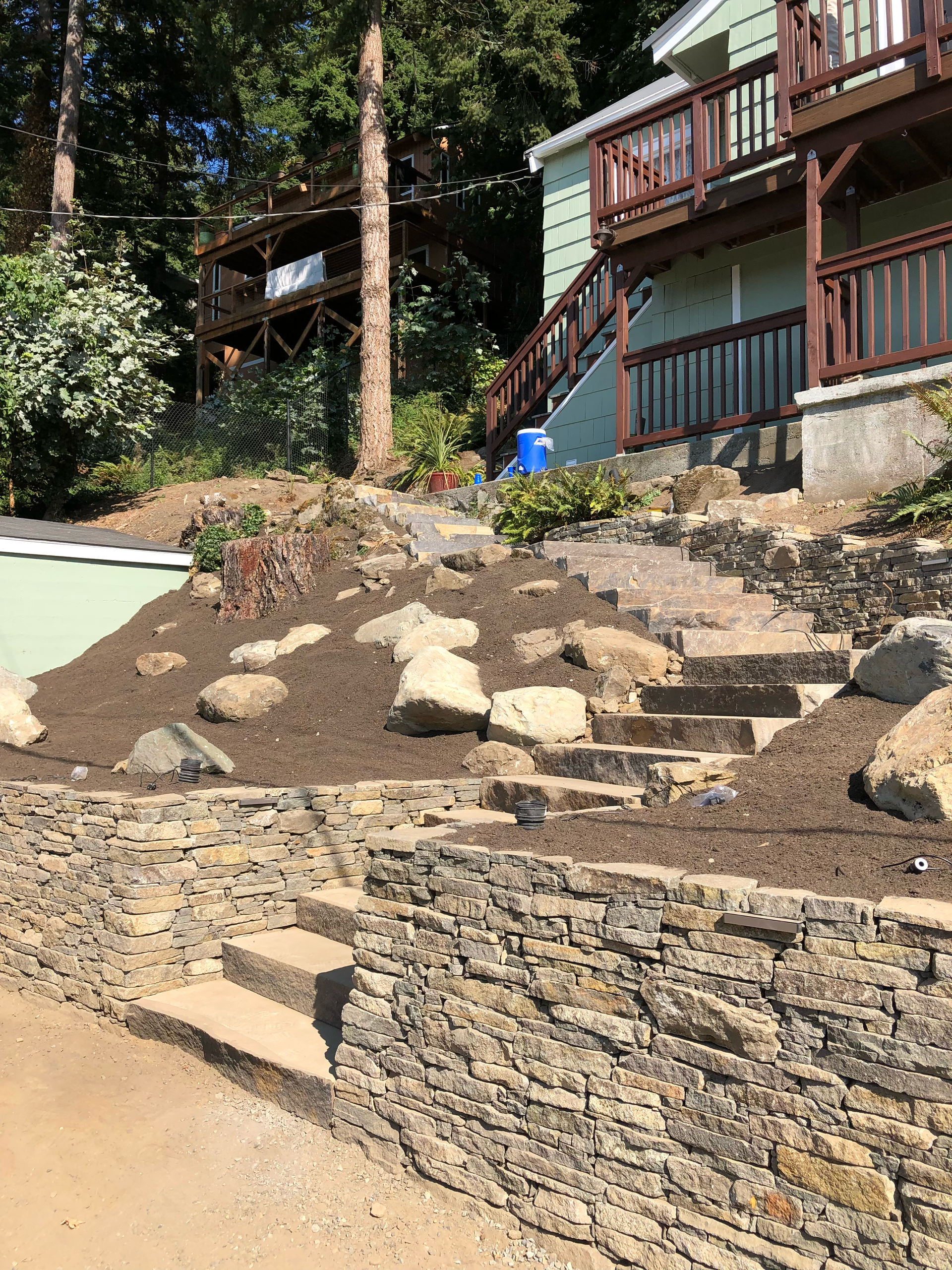 Stone steps and retaining walls ascend a hillside to a light green house with wooden decks; work by West Coast Landscape & Hardscape. 
