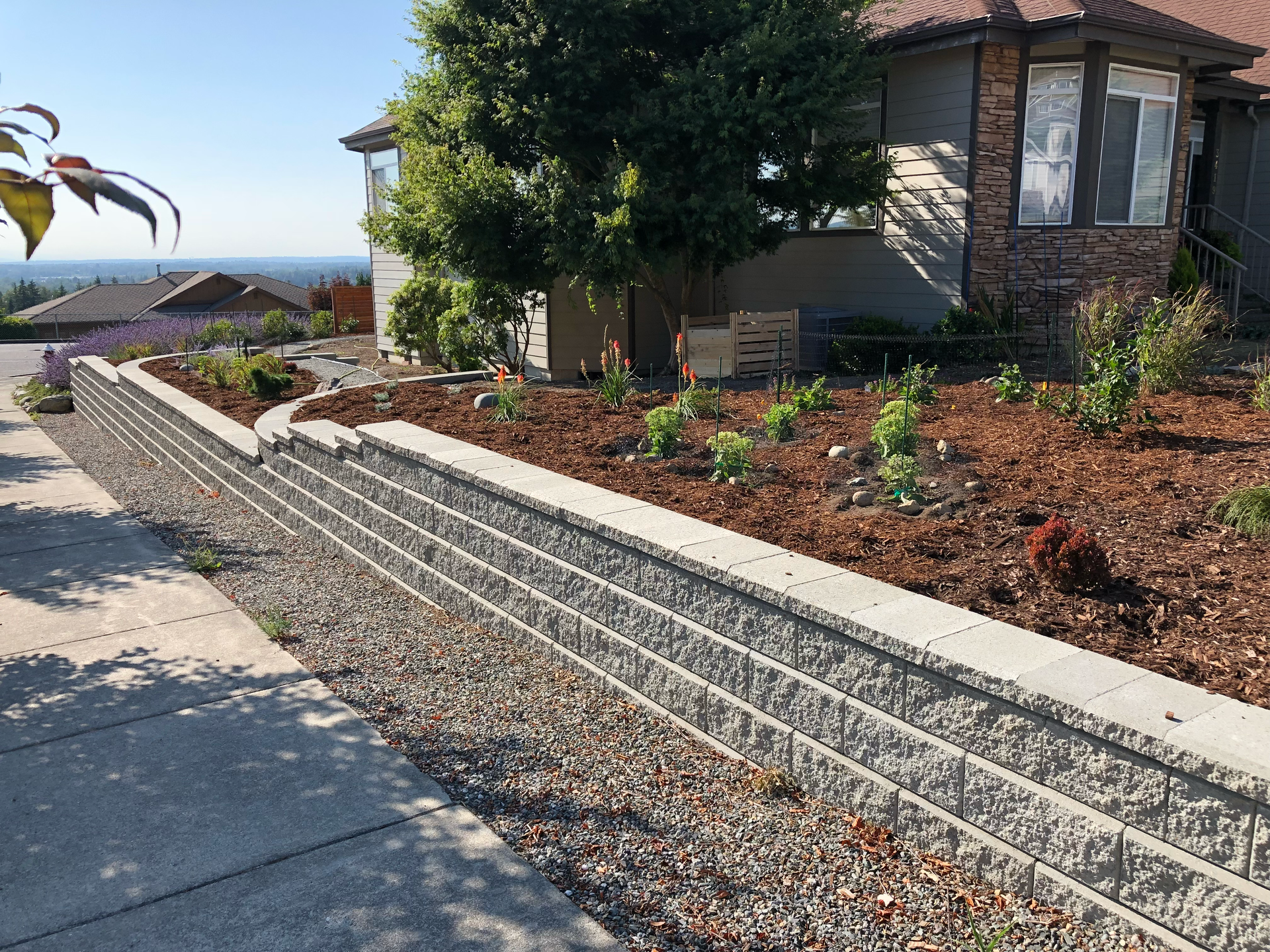 Stone retaining wall with a landscaped garden in front of a house, ocean visible in ; work by West Coast Landscape & Hardscape. 