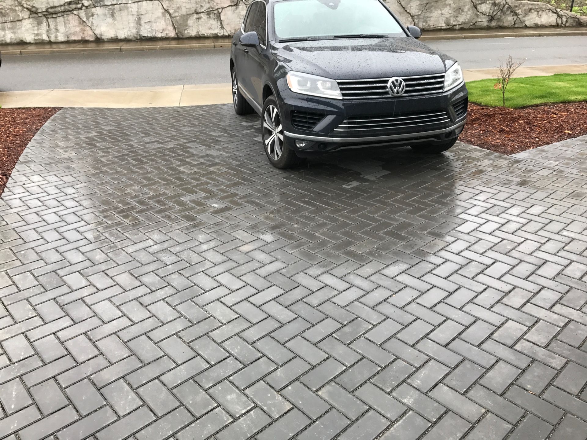 Dark SUV parked on wet, gray brick driveway with herringbone pattern; work by West Coast Landscape & Hardscape. 