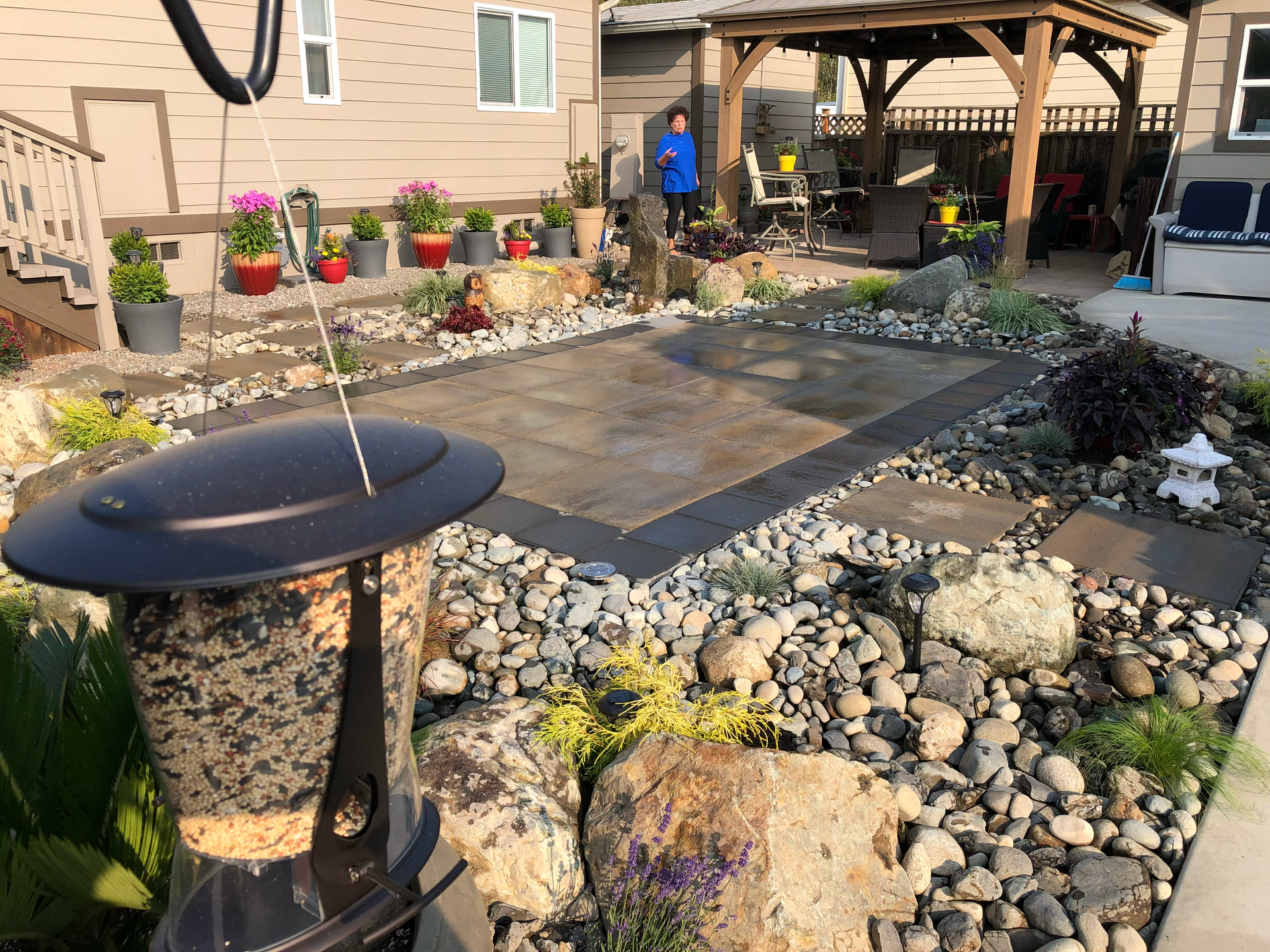 Backyard with a bird feeder, a covered rectangular structure, rocks, plants, and a person standing under a pergola; work by West Coast Landscape & Hardscape. 