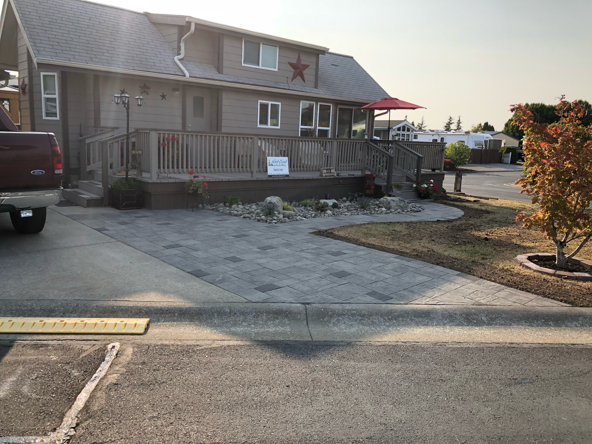 Two-story building with deck and red star. Gravel driveway with curb. Red truck parked; work by West Coast Landscape & Hardscape. 