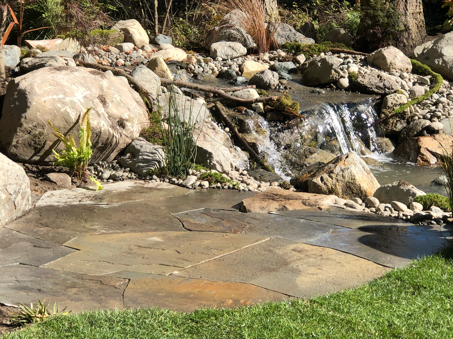Stone patio with a small waterfall feature and rocks in a garden setting; work by West Coast Landscape & Hardscape. 