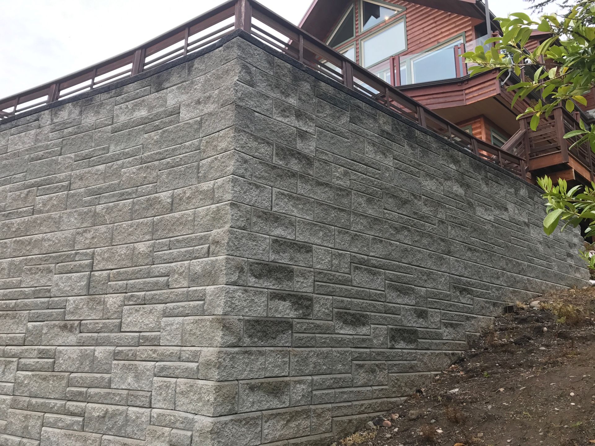 Gray stone retaining wall supporting a house with a wooden balcony and railing; work by West Coast Landscape & Hardscape. 