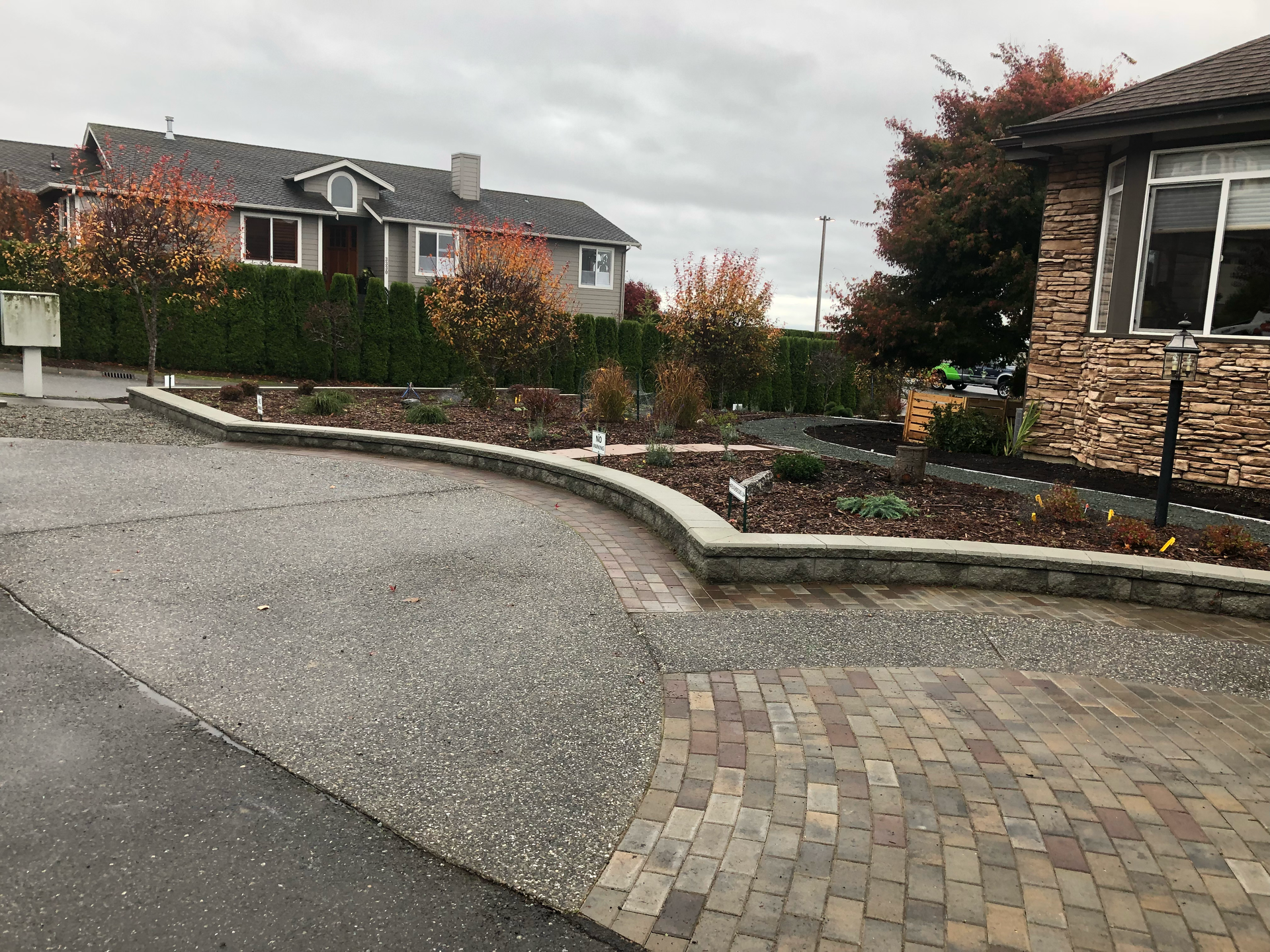 Curved driveway with brick and textured stone, bordered by a garden bed with colorful fall foliage. Houses in background; work by West Coast Landscape & Hardscape. 