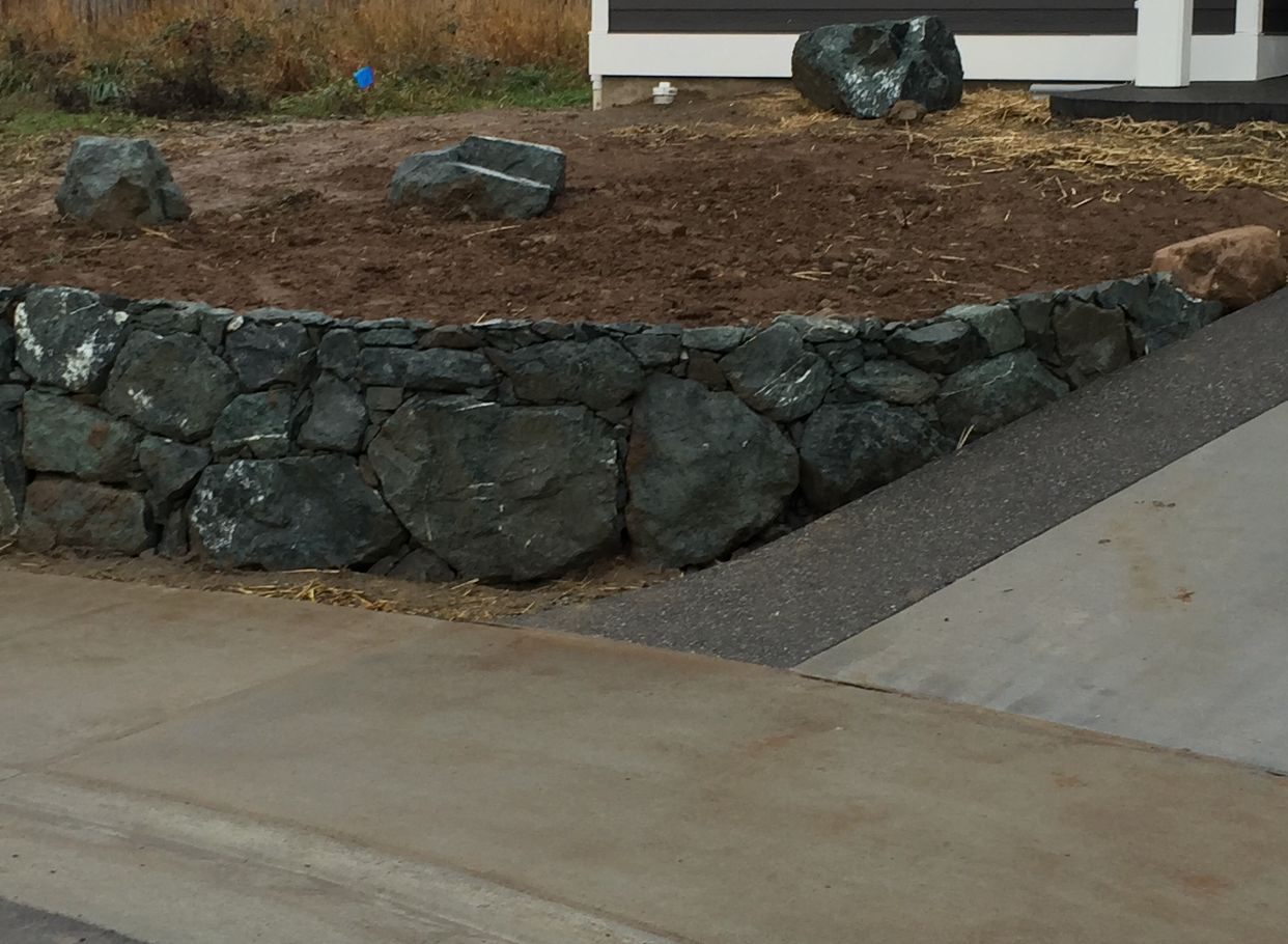 Stone retaining wall with large boulders, soil, and gravel, next to a concrete driveway; work by West Coast Landscape & Hardscape. 