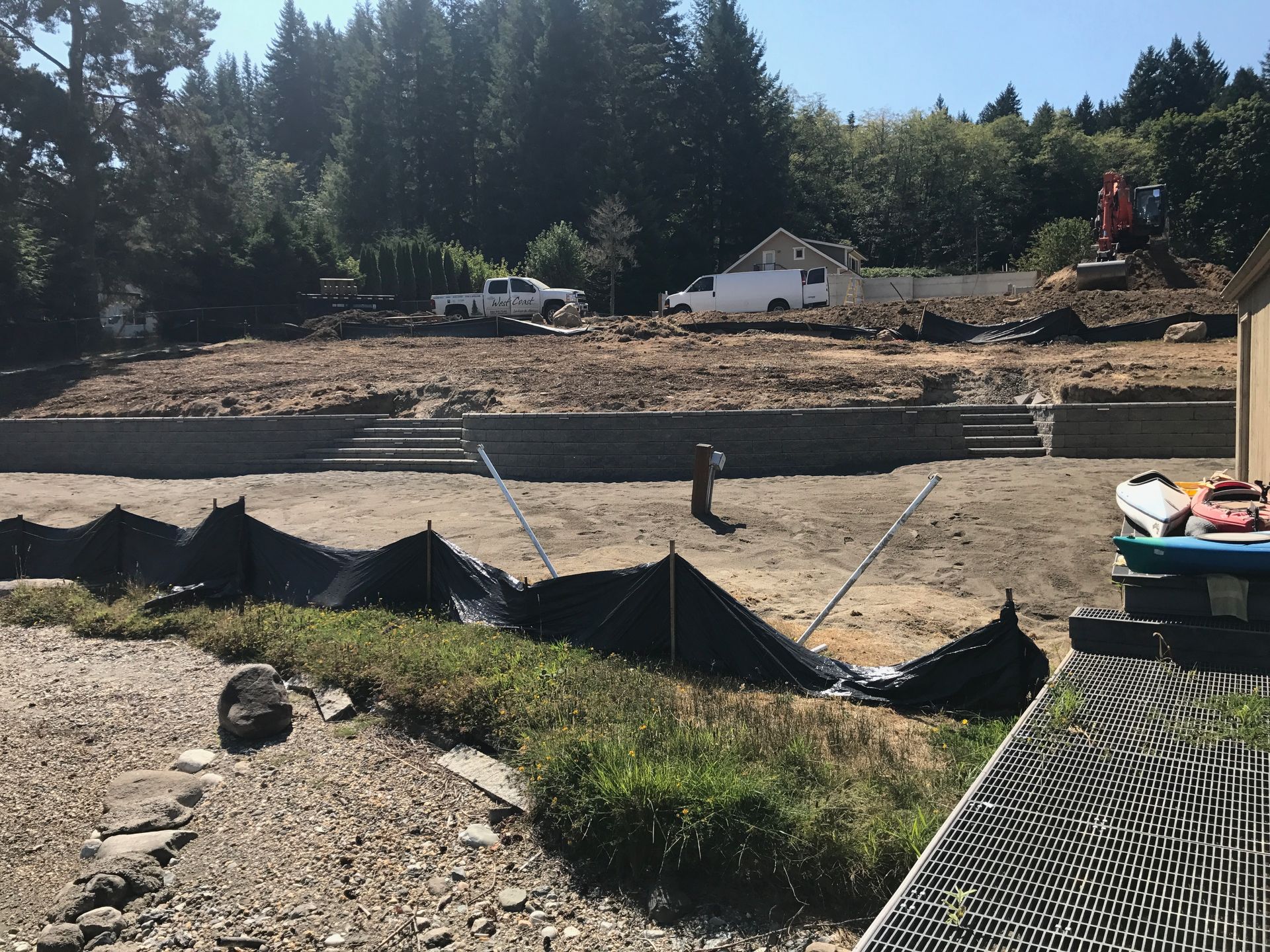 Construction site near water with soil, barriers, trucks, and trees in background; work by West Coast Landscape & Hardscape. 