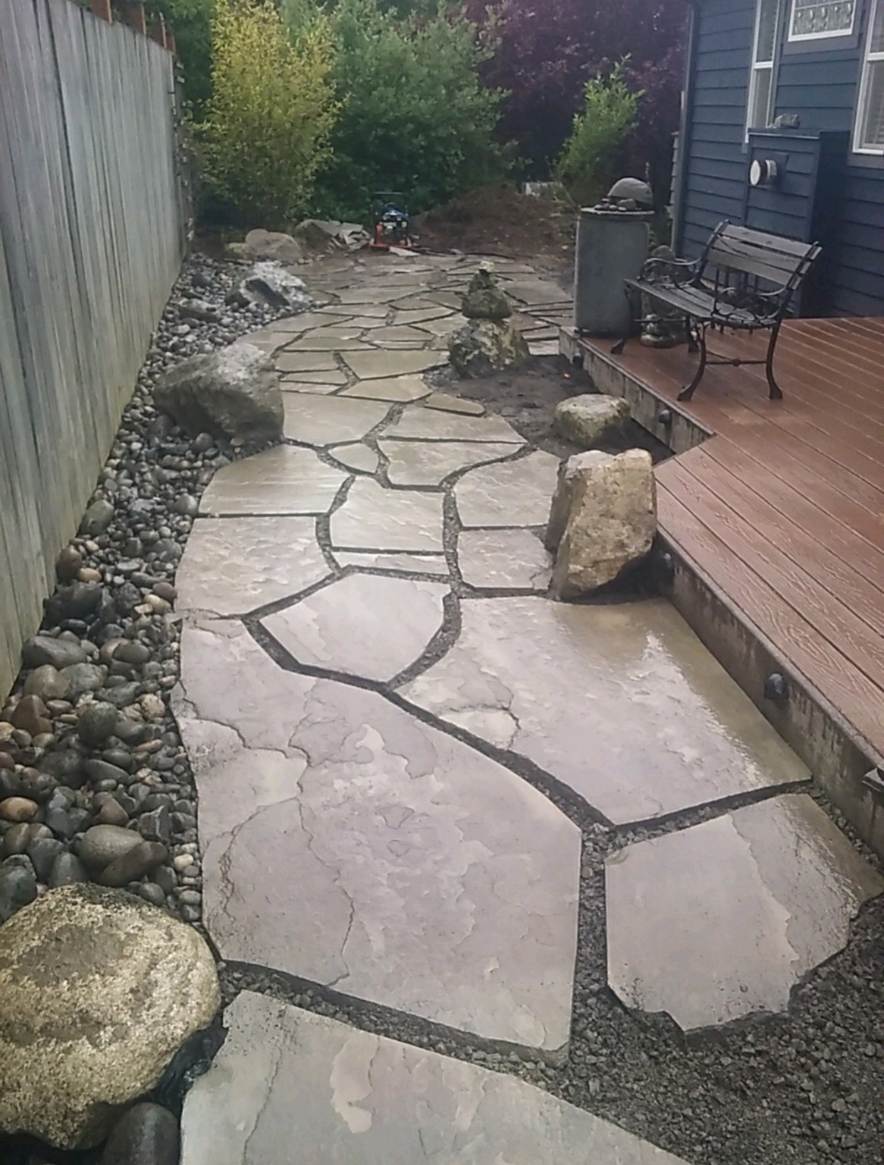 Flagstone path with large rocks, leading from deck with bench to a garden with fence; work by West Coast Landscape & Hardscape. 