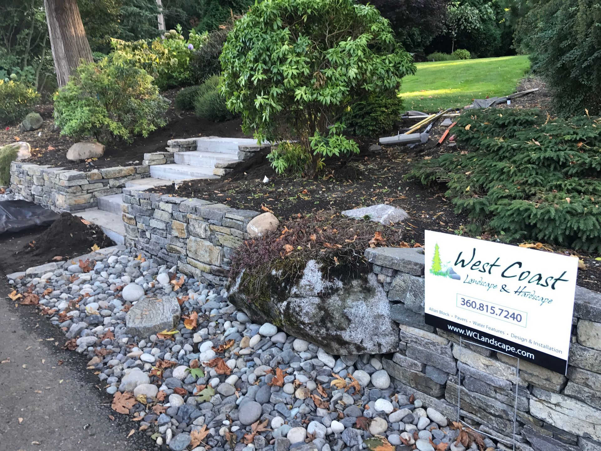 Stone retaining wall and steps leading uphill, with landscaping and a sign for West Coast Landscape & Hardscape. 