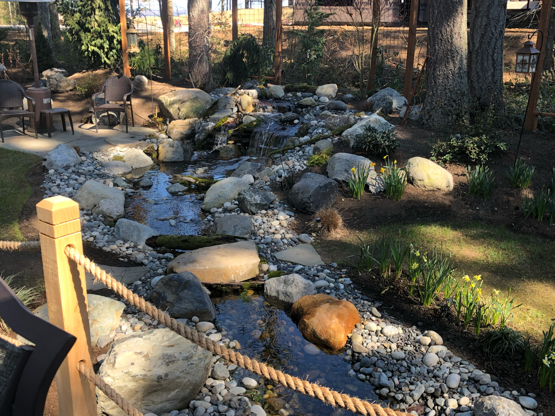 A small stream flows through a garden, surrounded by rocks and vegetation, with a rope fence; work by West Coast Landscape & Hardscape. 