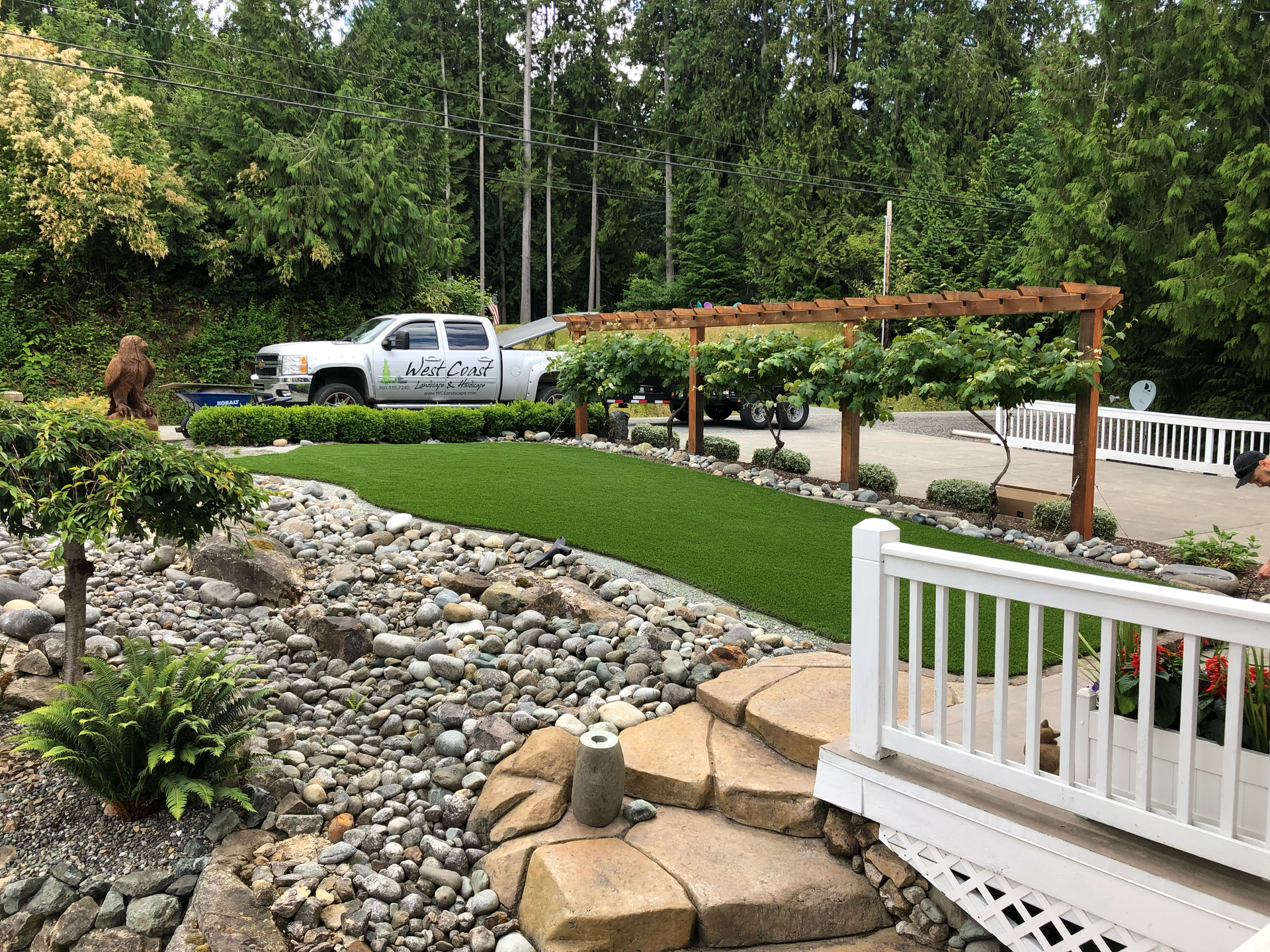 Landscaped yard with stone pathway, artificial turf, wooden pergola, and white pickup truck; work by West Coast Landscape & Hardscape. 